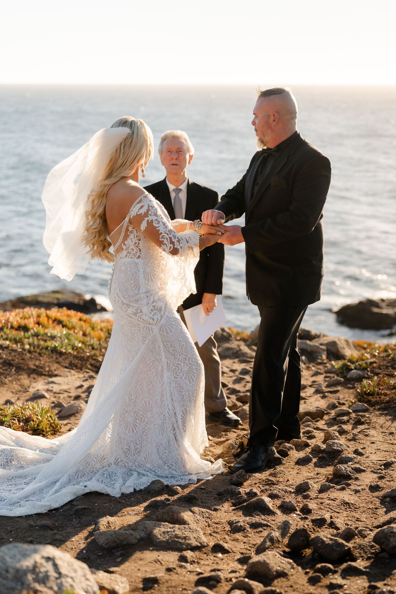 Wedding couple dances together on a scenic beach with rocks and ocean waves in the background.