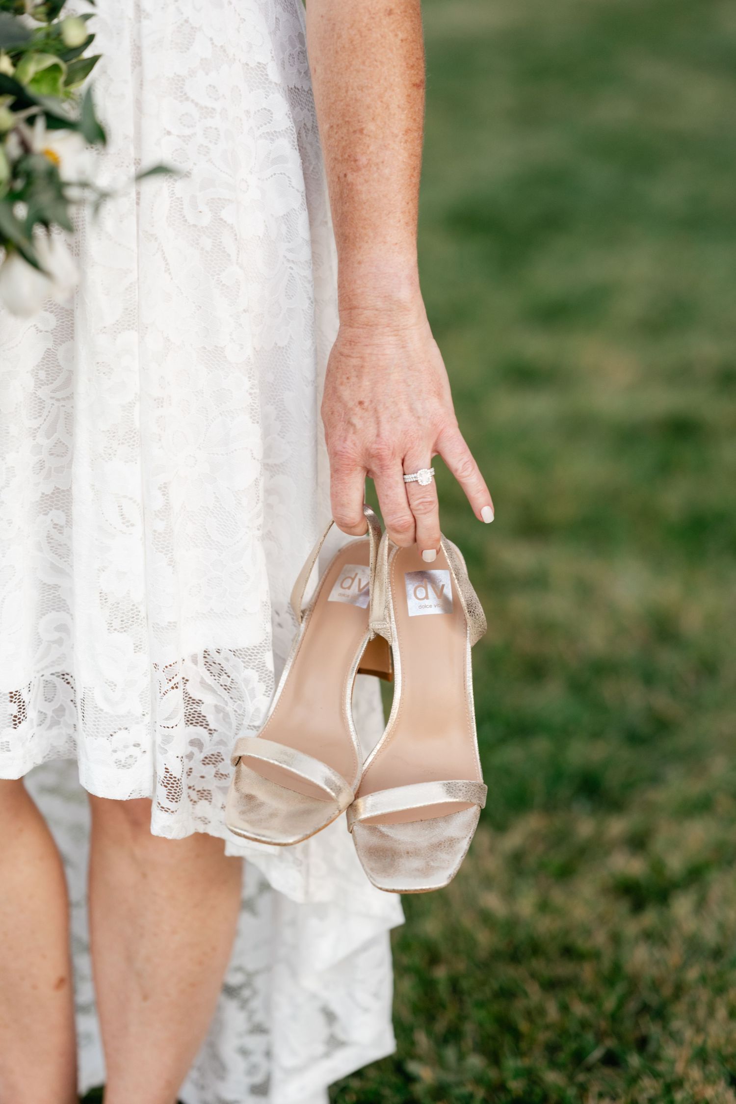 A close-up of a bride holding nude wedding sandals against a lace dress hem on grass.