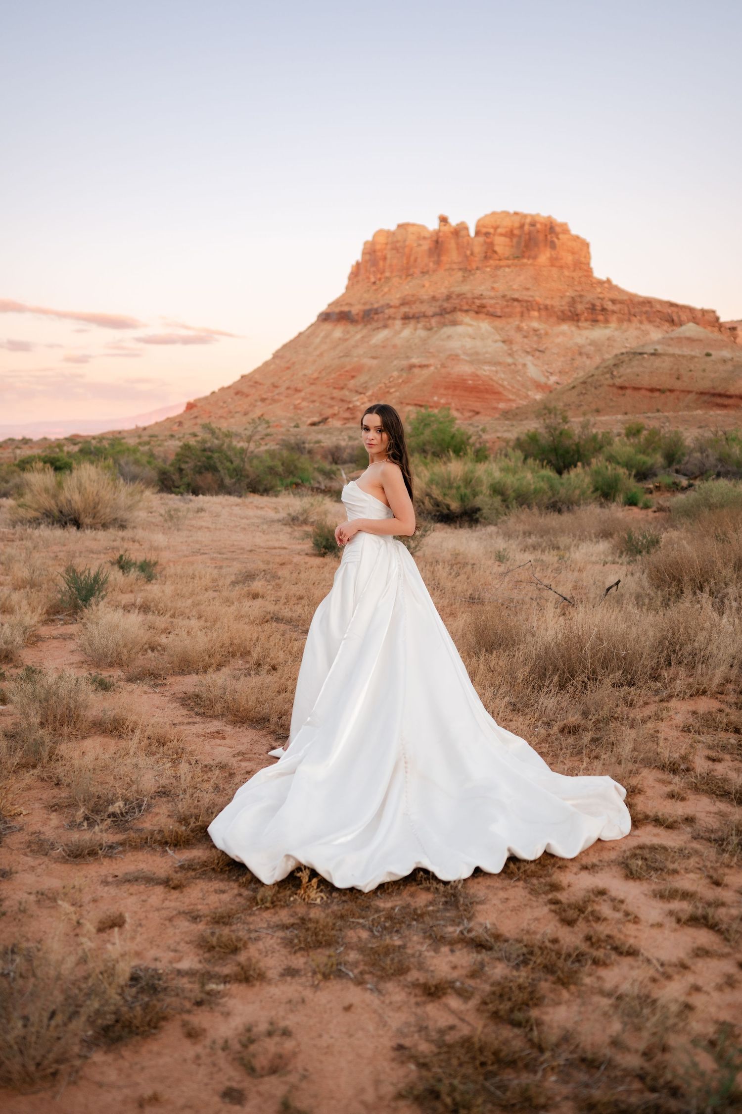 Bride in flowing white gown poses before red rock mesa at sunset in desert landscape.