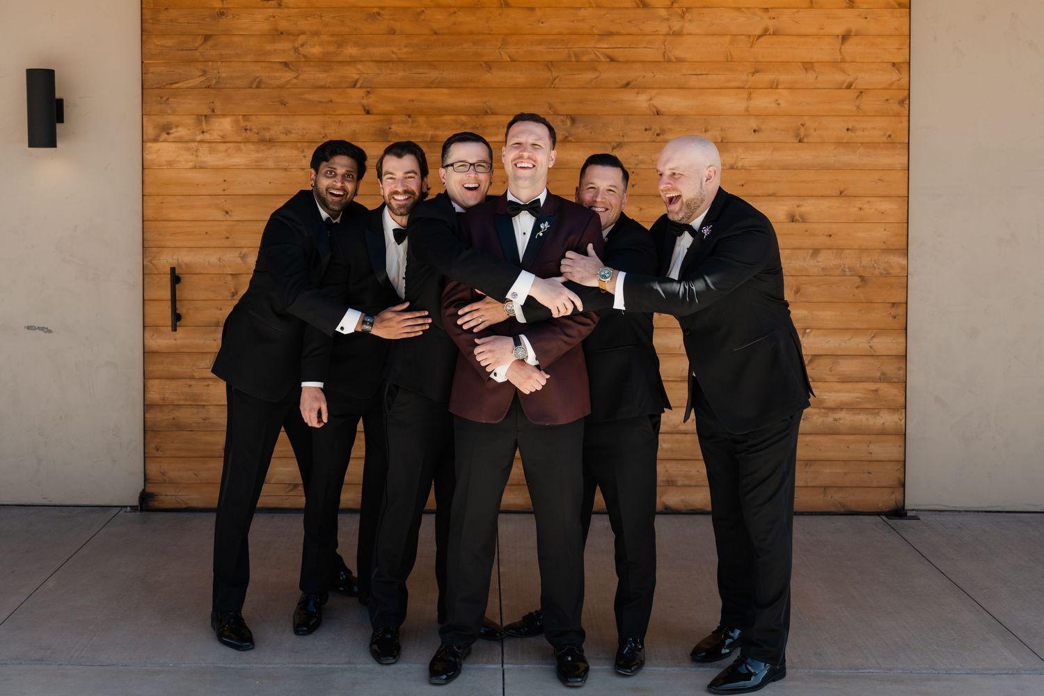 Groom in burgundy tuxedo surrounded by smiling groomsmen in black suits posing playfully at The Red Earth Venue in Moab, Utah.