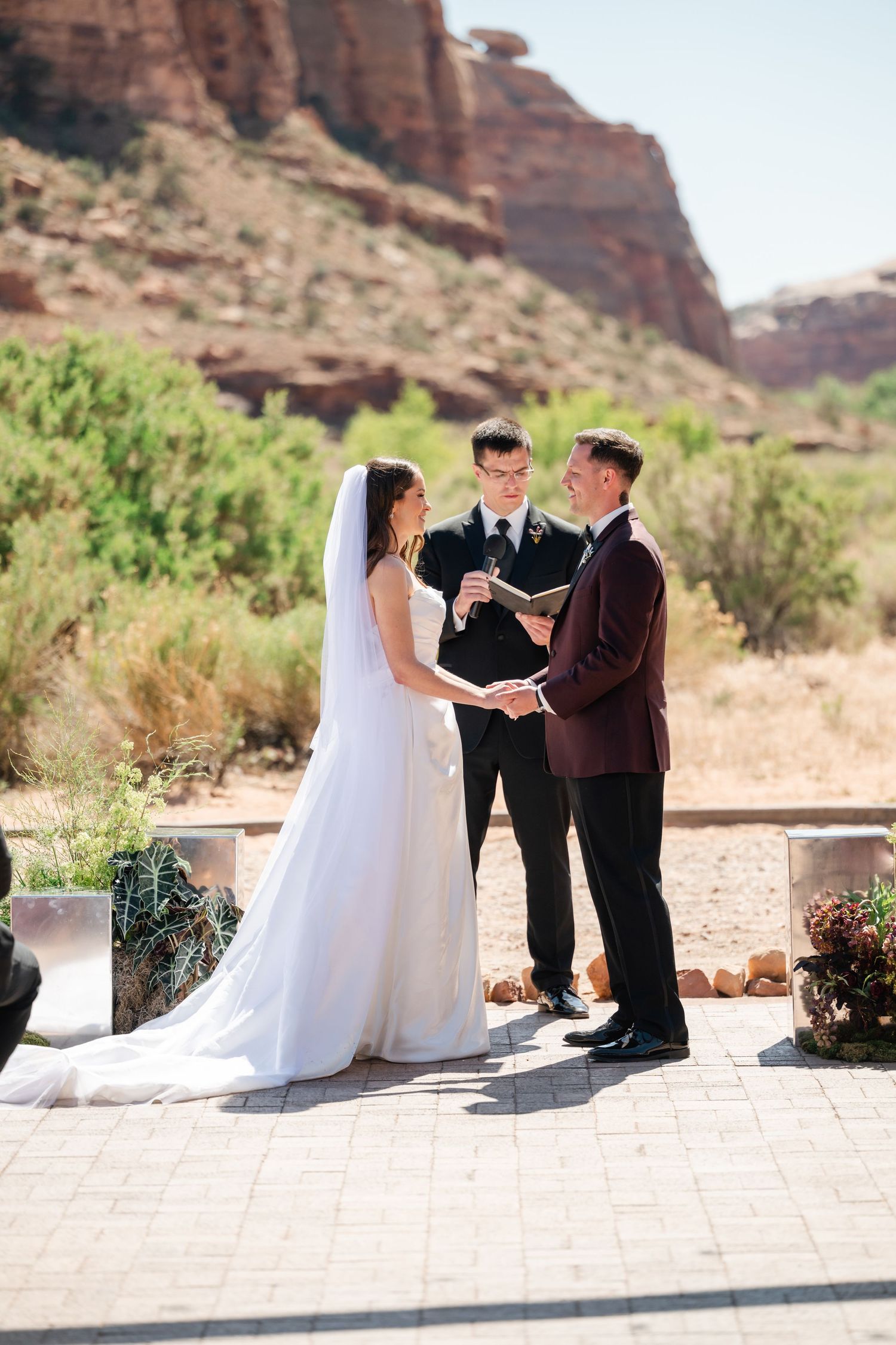 Bride and groom exchange vows outdoors with an officiant, red rock canyon landscape behind them on a sunny day.