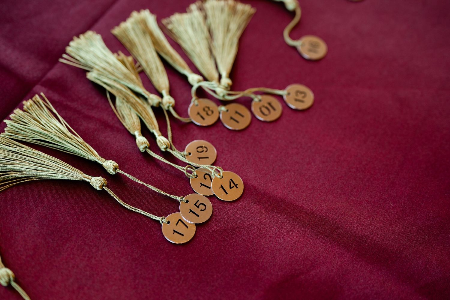 Gold tassel bookmarks with numbered copper tags arranged on a rich burgundy red fabric background.