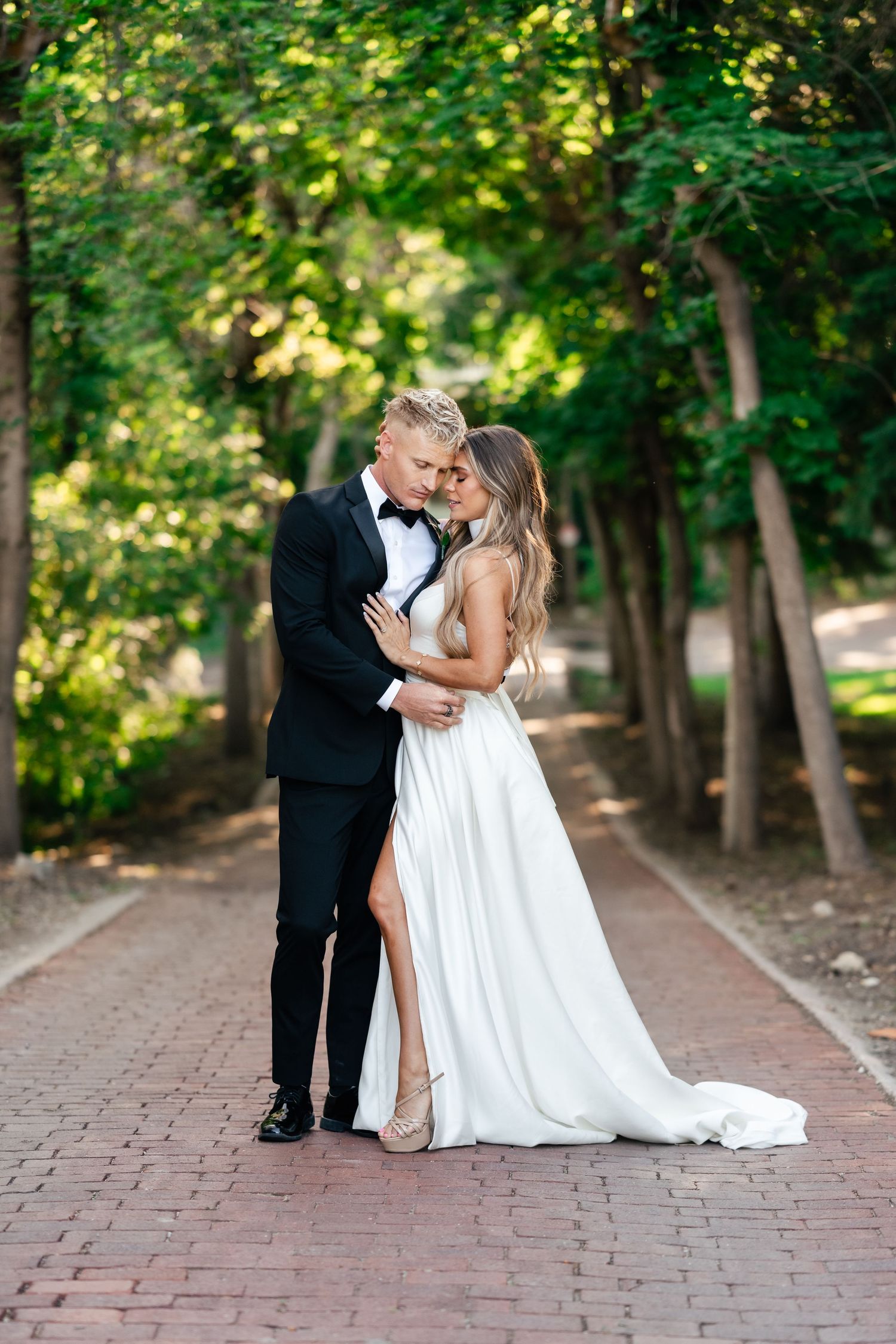Elegant couple embraces on a brick pathway surrounded by lush green trees in romantic wedding attire.