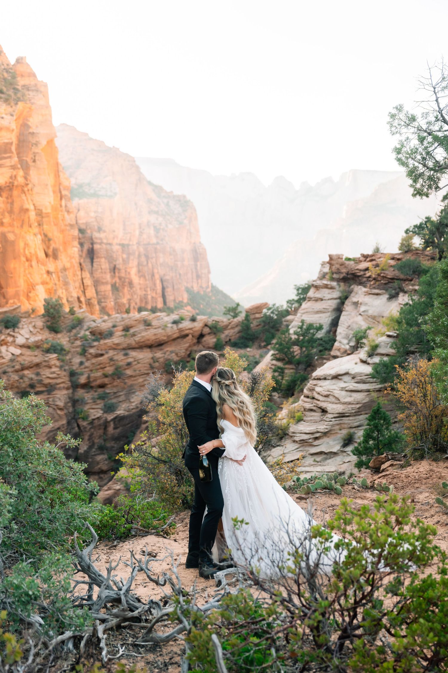 Wedding couple embracing on desert trail with towering sandstone cliffs in background.