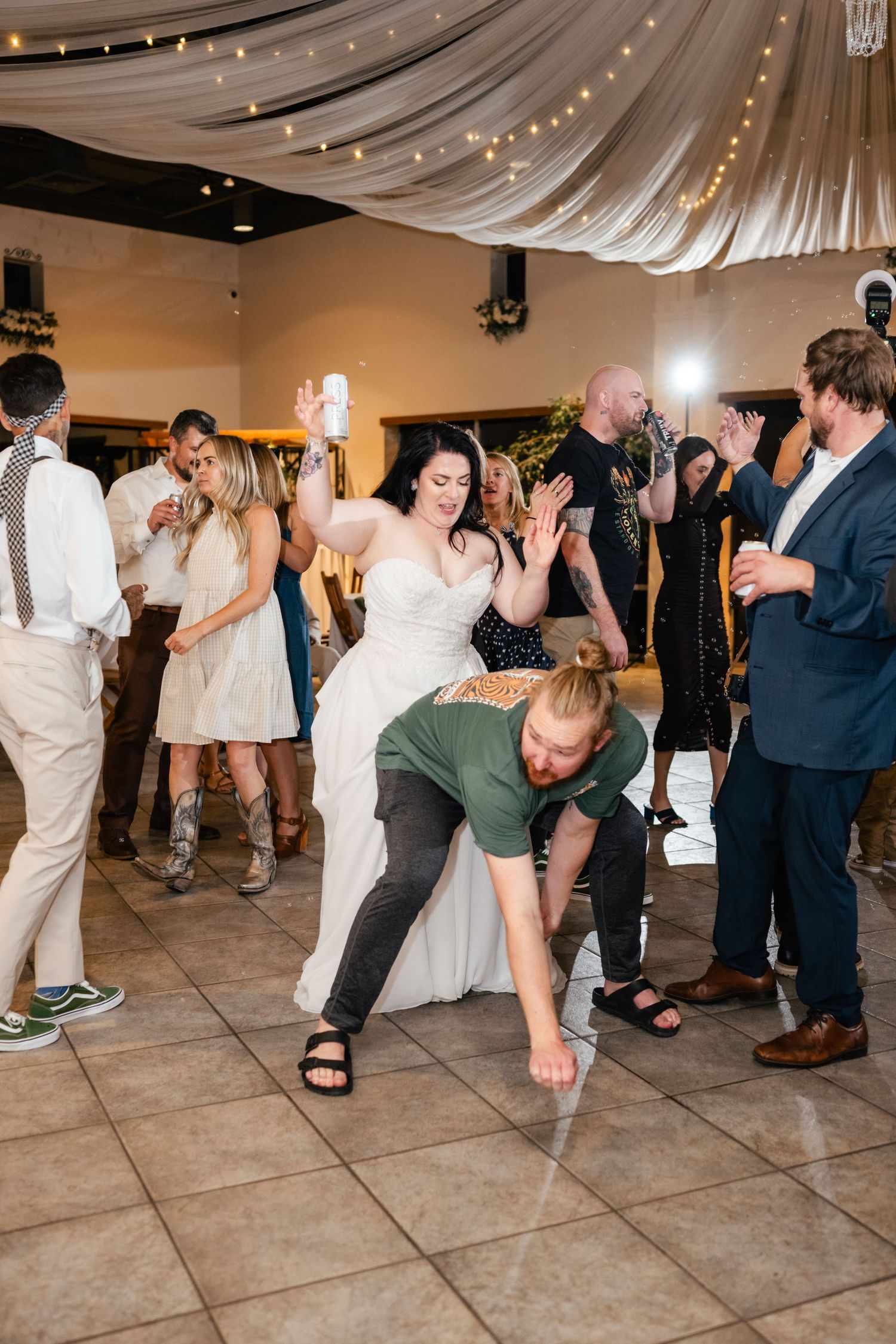 Wedding reception dance party shows guests enjoying themselves on a ballroom floor under draped fabric and string lights.