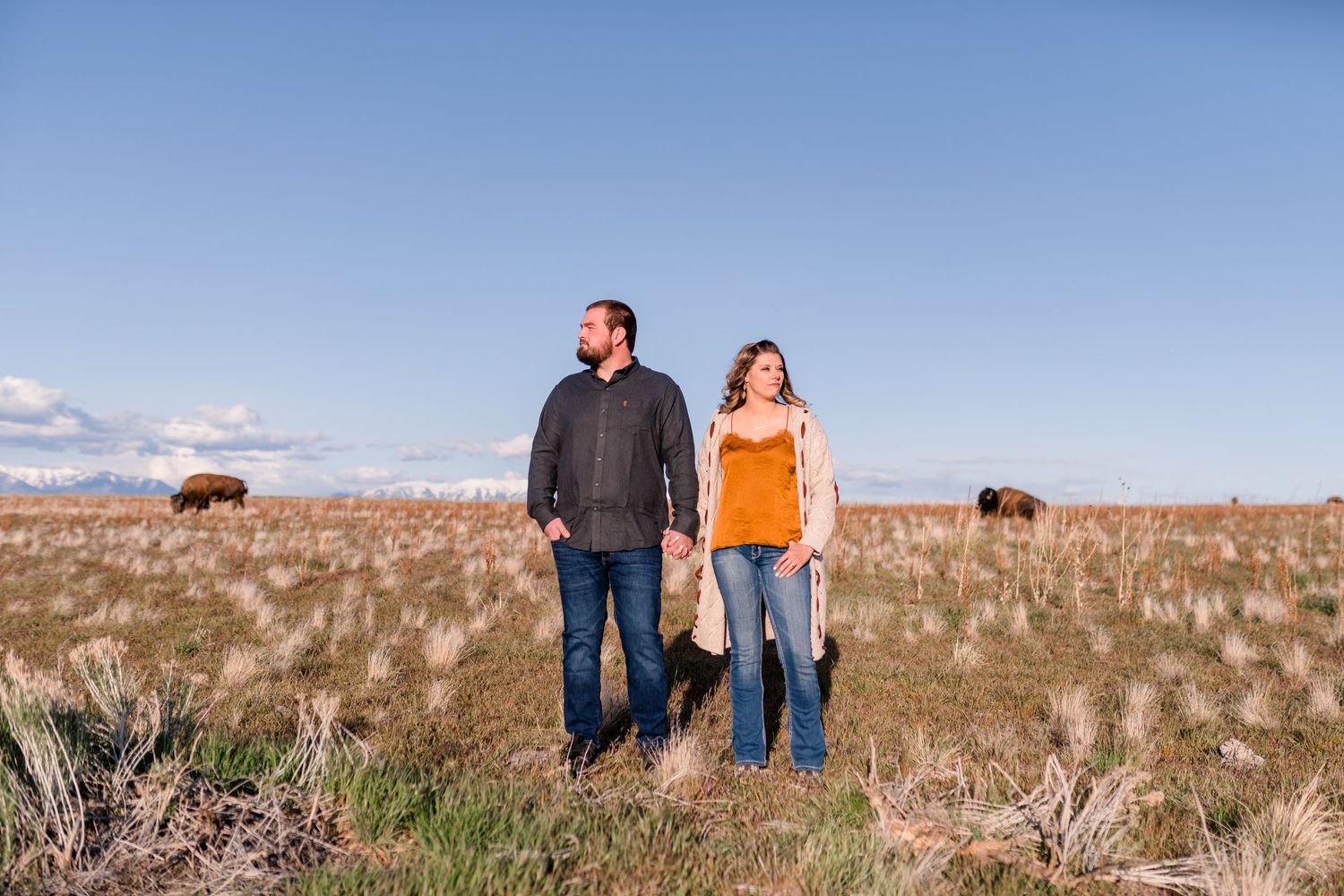 A couple standing together in a rustic field with grazing bison in the background.