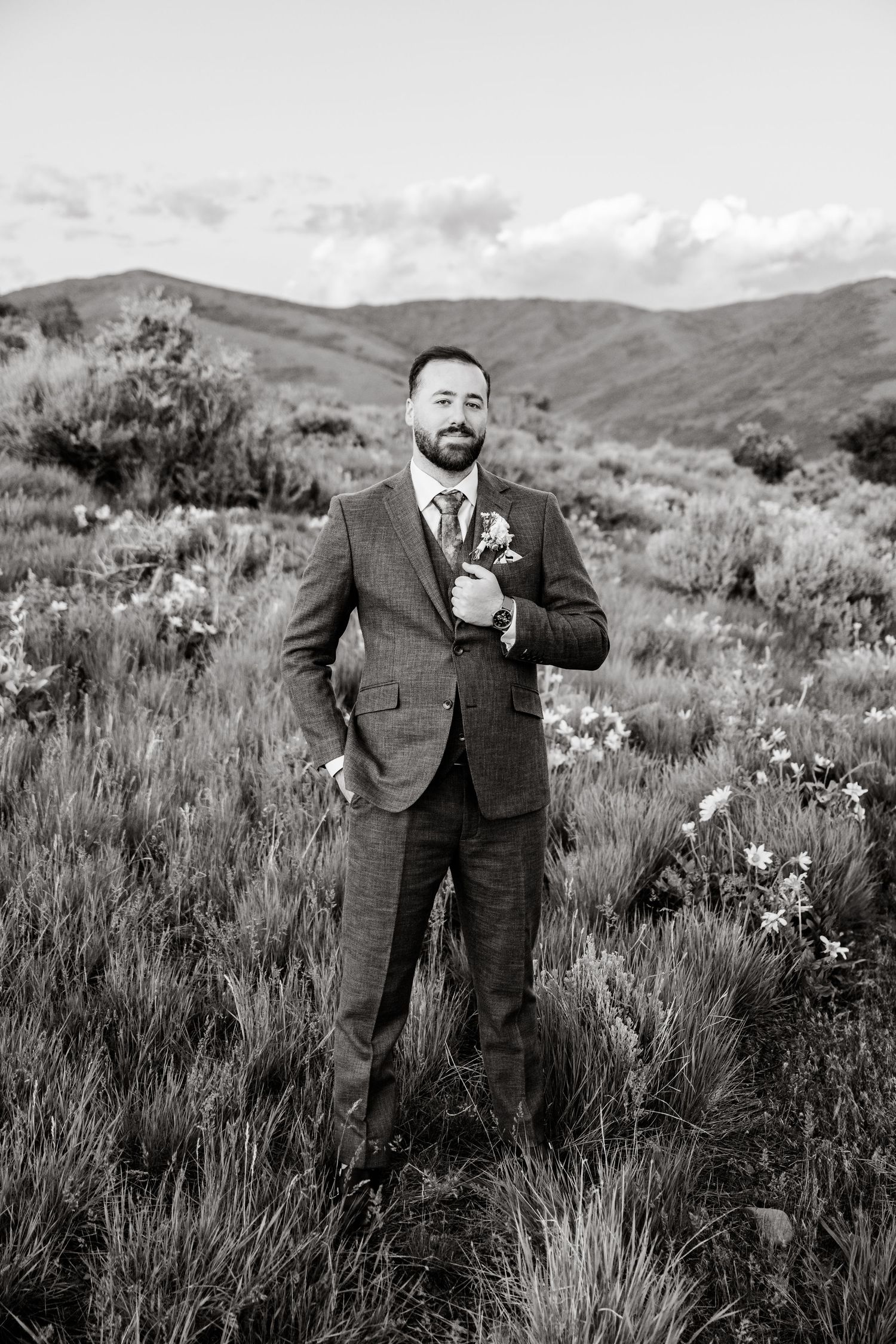 Groom in charcoal suit poses among sage brush with rolling hills in black and white photography.