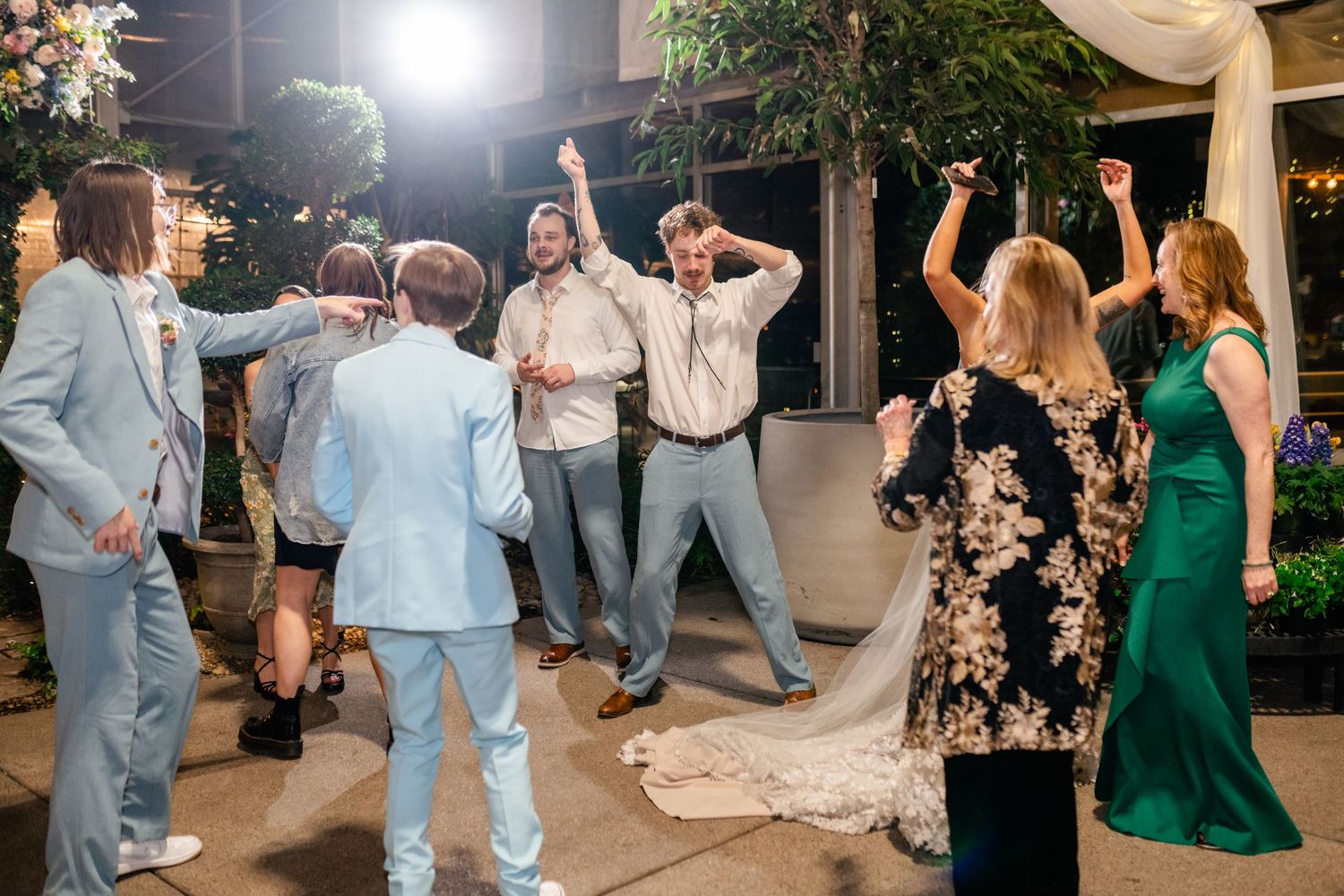Wedding guests dance and celebrate on an outdoor patio at night under string lights at a reception.