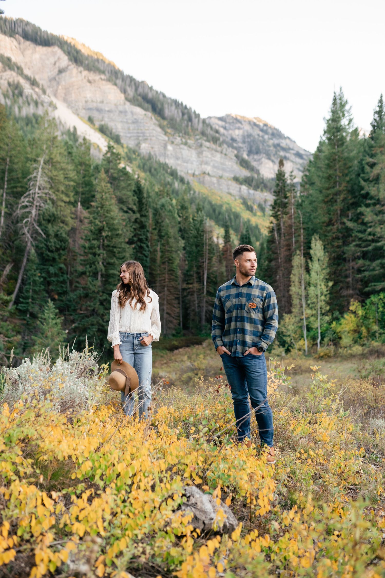 A couple explores a mountain trail surrounded by yellow wildflowers and towering evergreen forests.