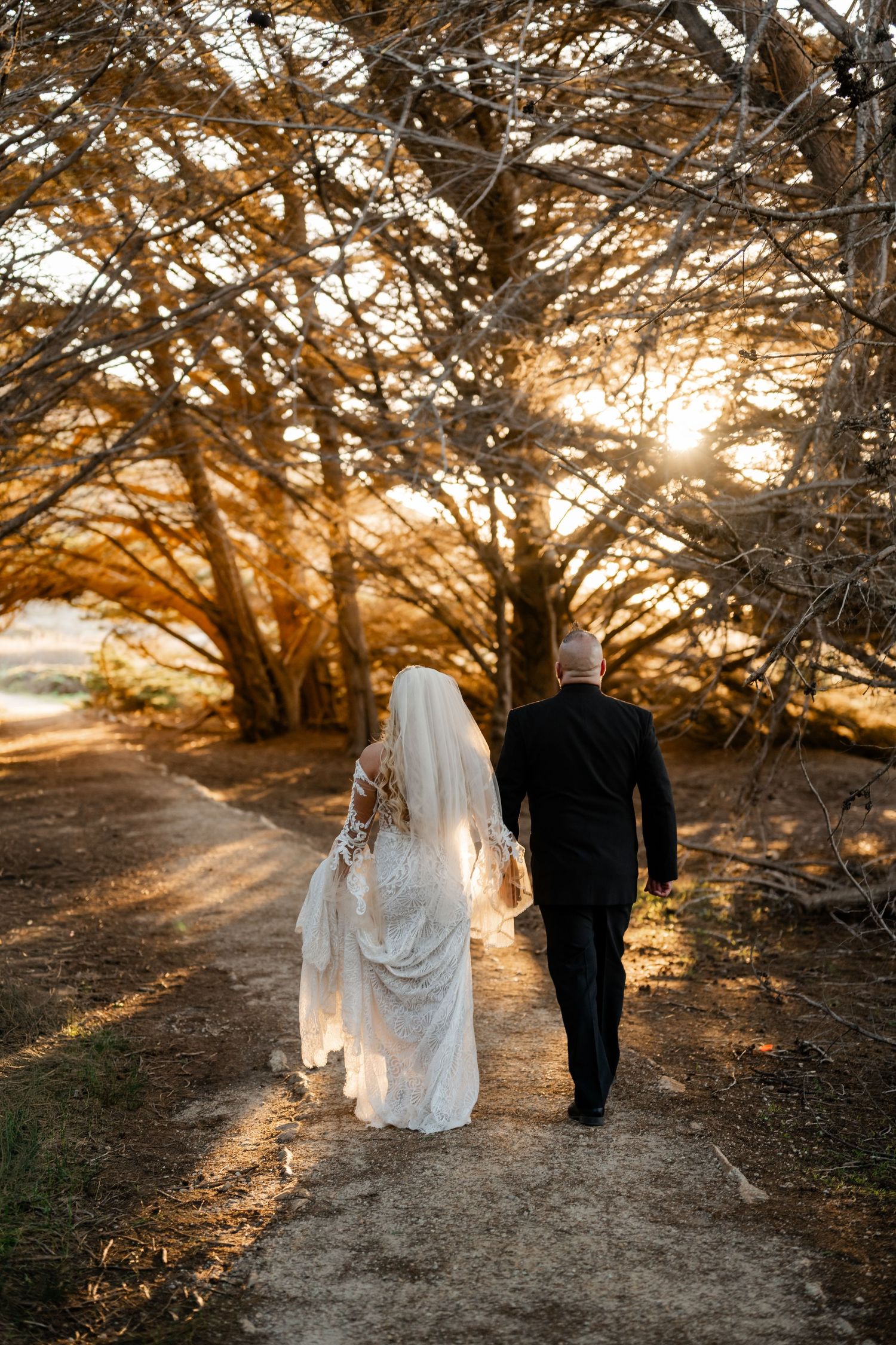 A couple walking down a sunlit path through trees during golden hour on their wedding day.