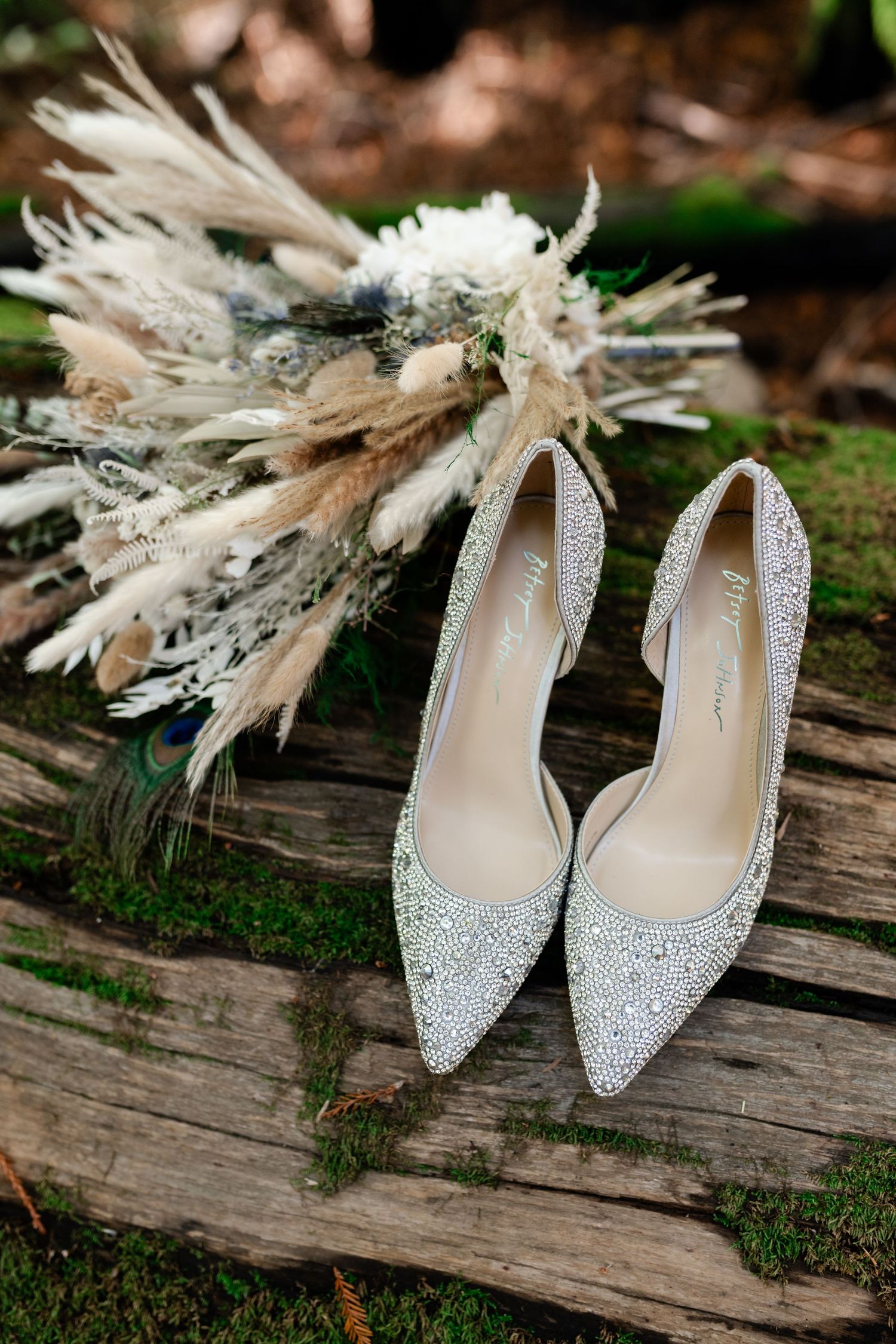 Elegant white bridal shoes with feather accessories arranged on weathered wood in natural outdoor setting.