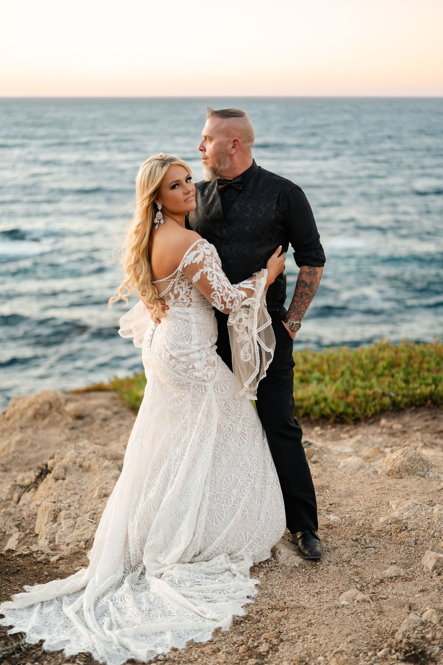 A couple embraces on a scenic coastal cliff at sunset with the bride wearing a flowing lace wedding dress.