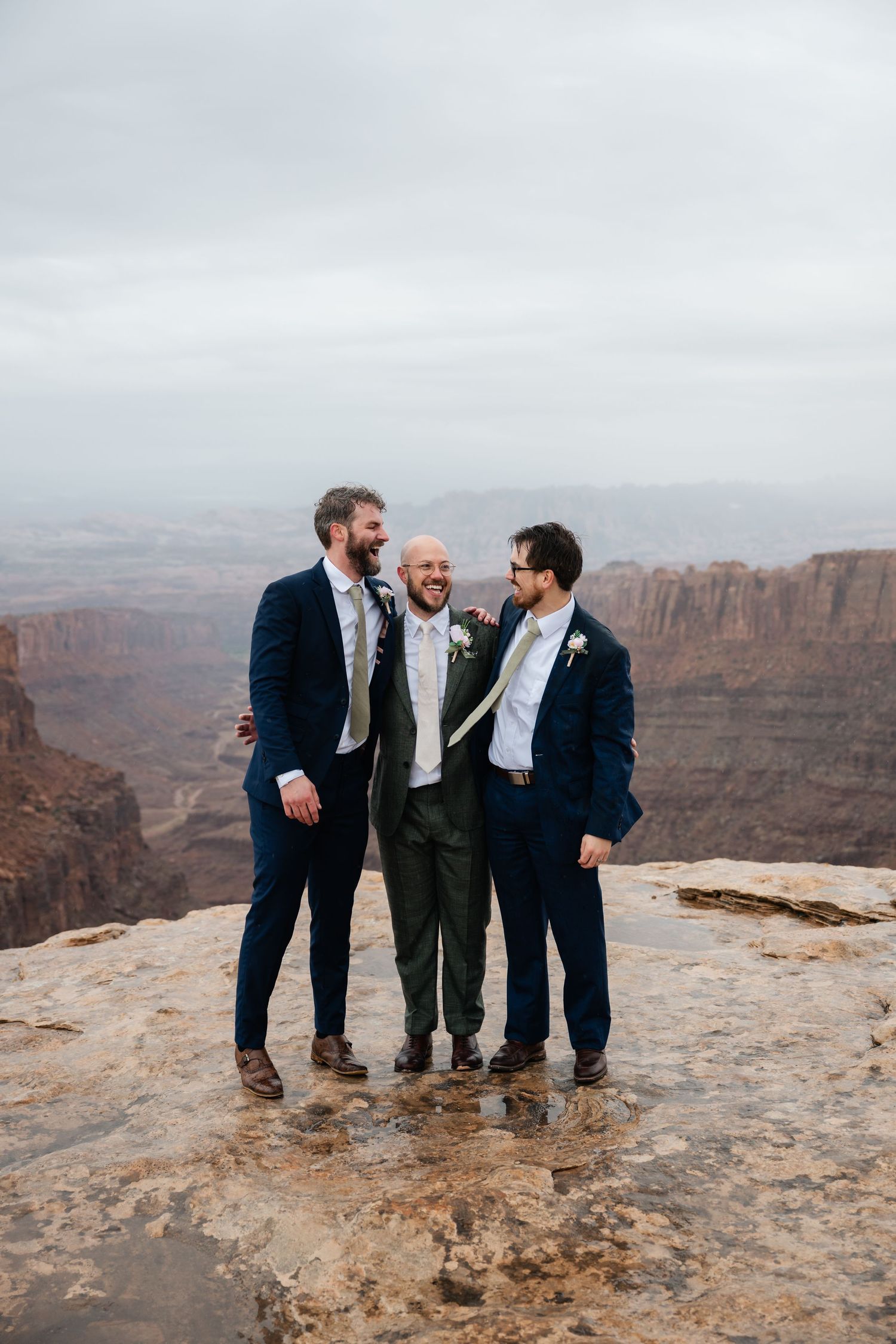 Three people in formal attire share a laugh while standing on rocky cliff edge with canyon views.