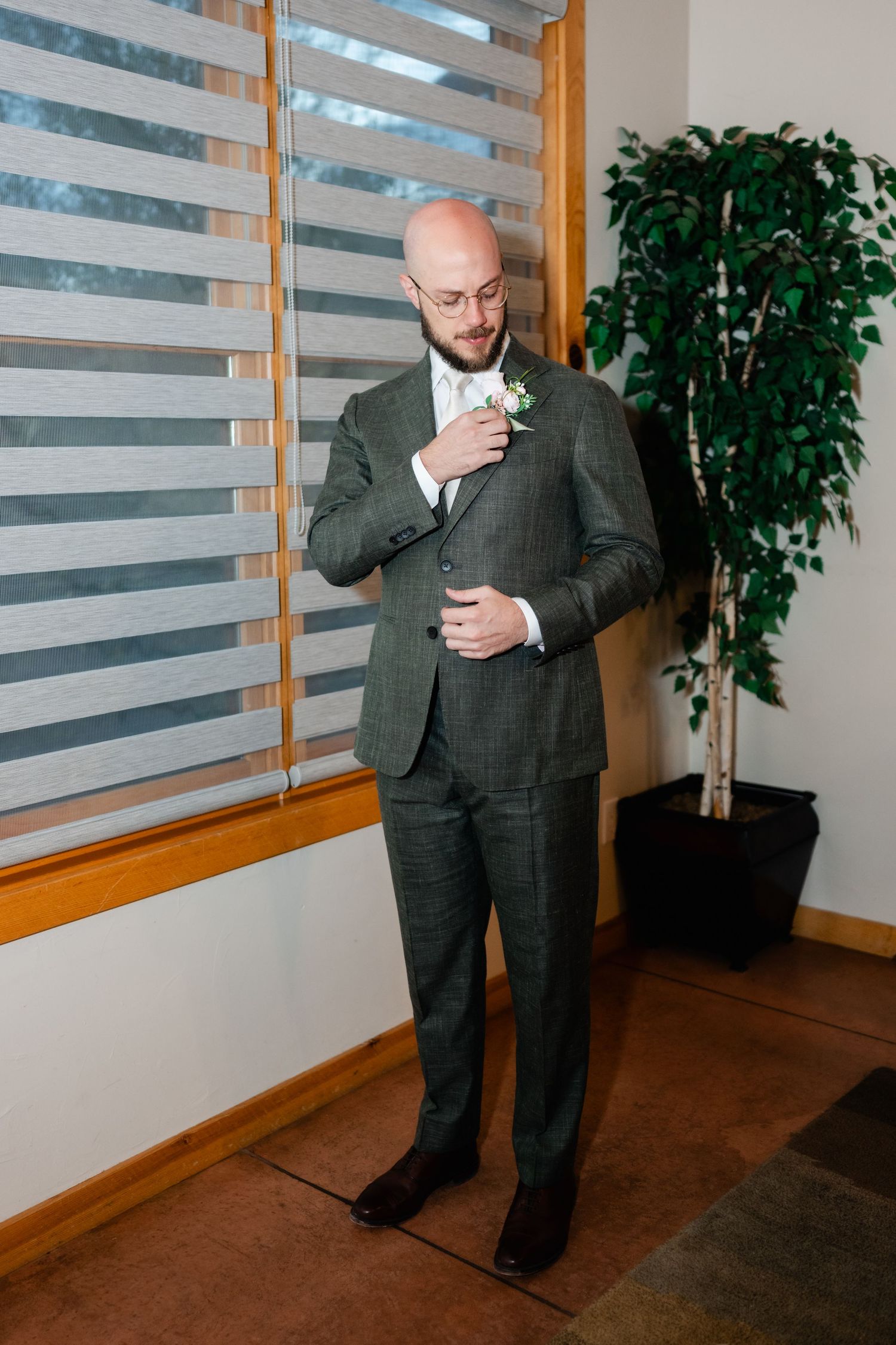 Groom in charcoal suit strikes poses in an indoor setting with window blinds and plants.