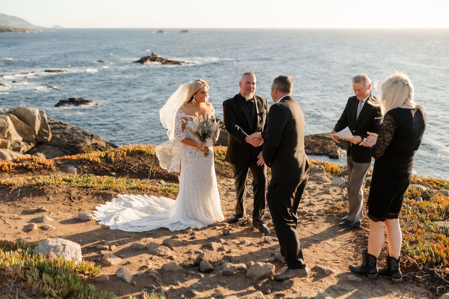 A romantic coastal wedding ceremony takes place on rugged cliffs overlooking the ocean at sunset.