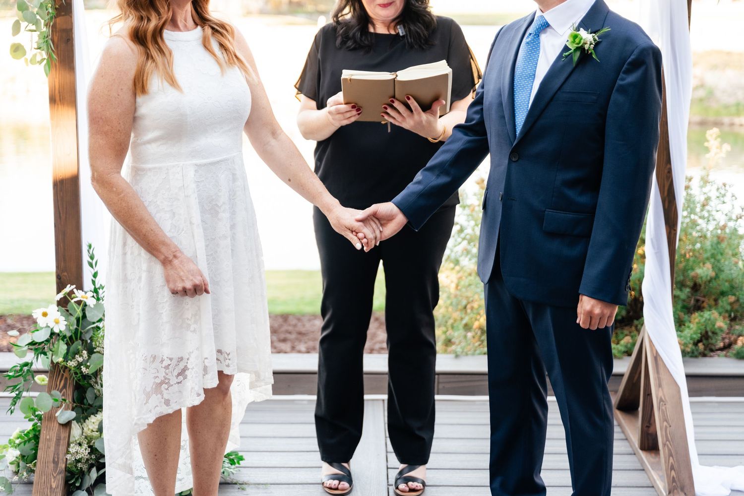 A couple stands at an outdoor wedding ceremony with an officiant reading from a book during their vows.