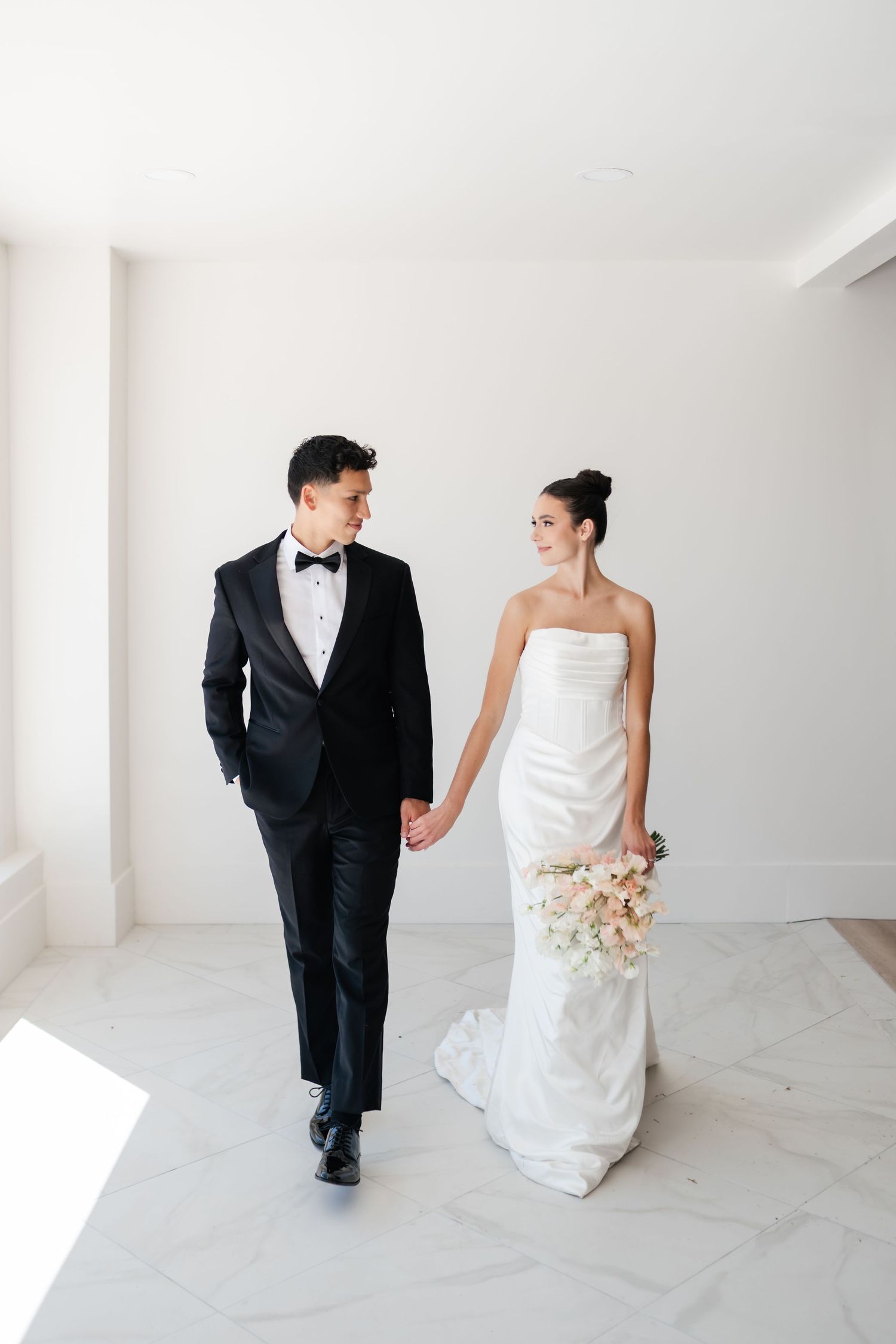 A bride and groom walk together in a minimalist white room during their wedding photo session.