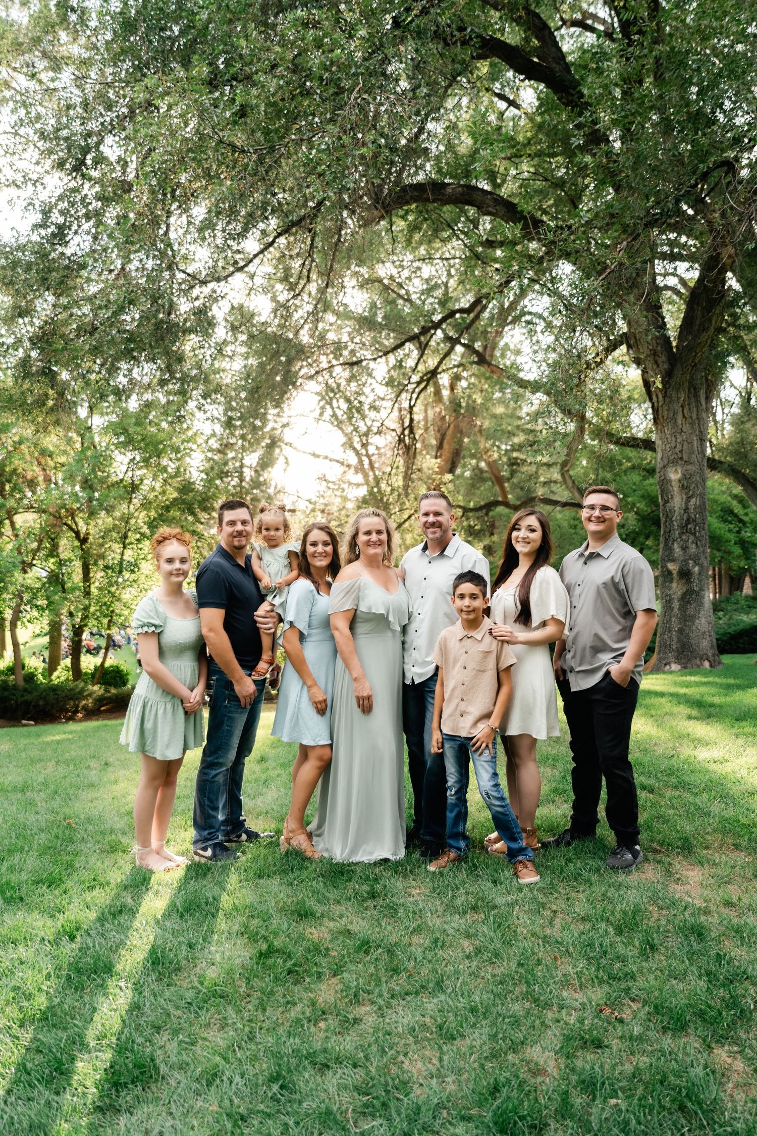 A group of people dressed in coordinating pastel and neutral tones pose together on a grassy lawn surrounded by trees.