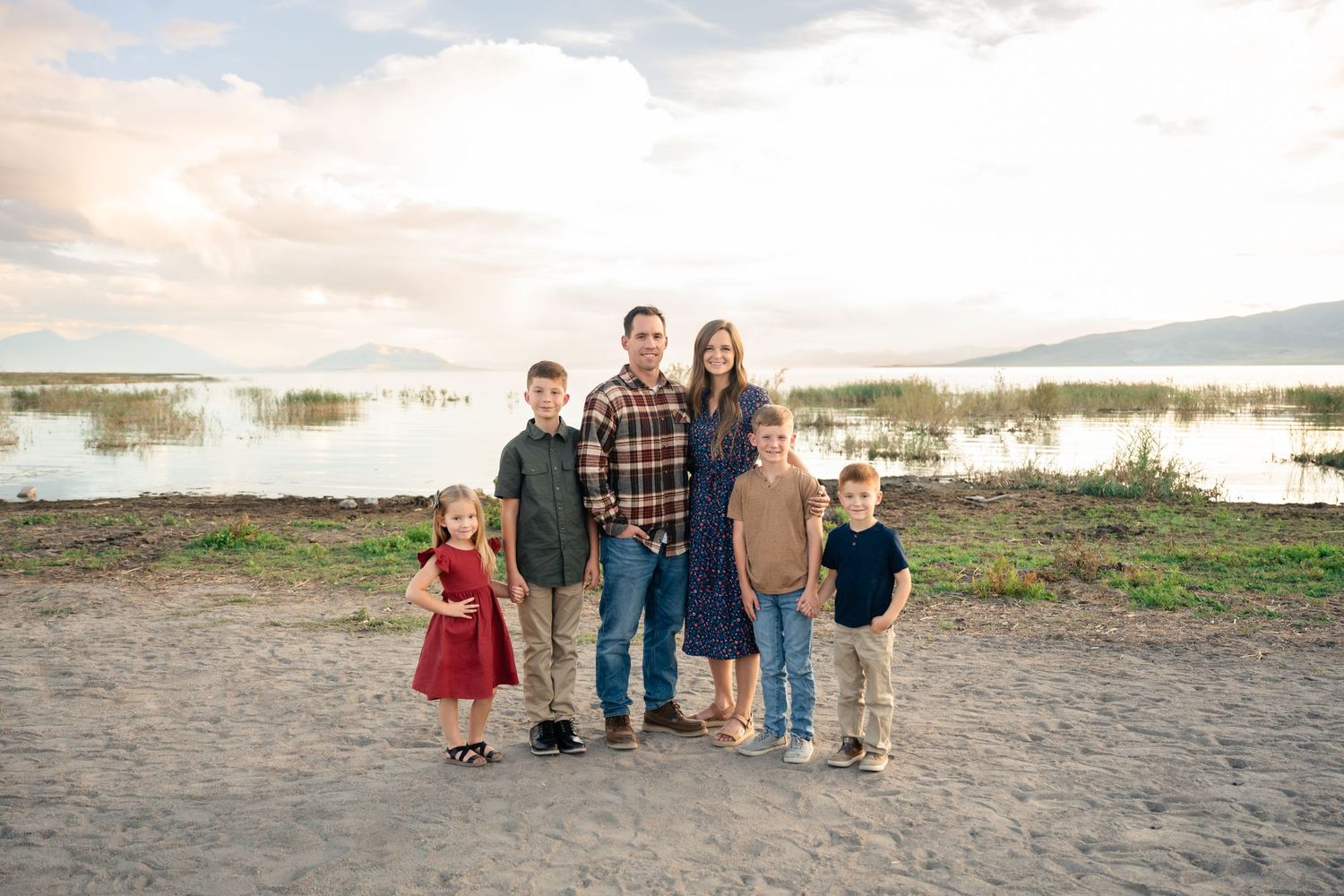 A group stands together outdoors at sunset near a lake with mountains visible in the background.