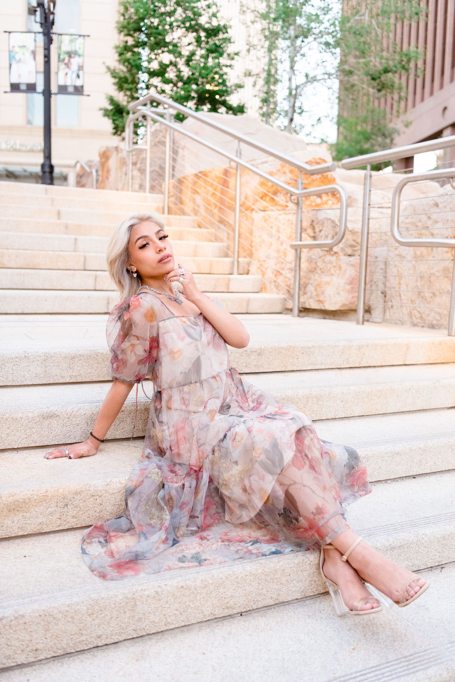 A person in a flowing floral dress poses gracefully on stone steps in an outdoor setting.