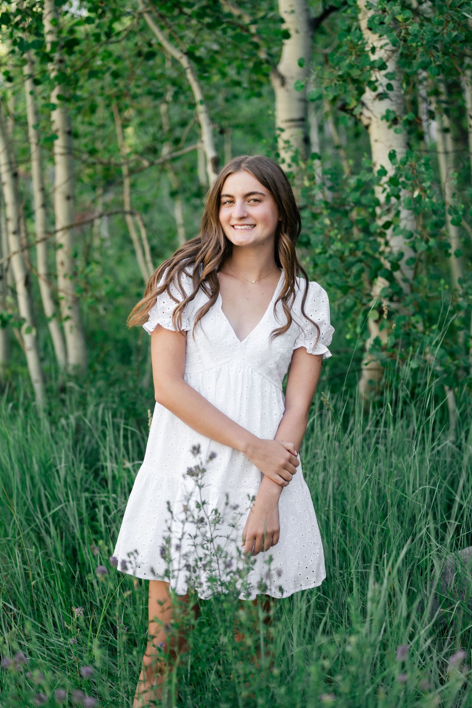 A person in a white dress poses gracefully among tall aspen trees and green grass in a summer forest setting.