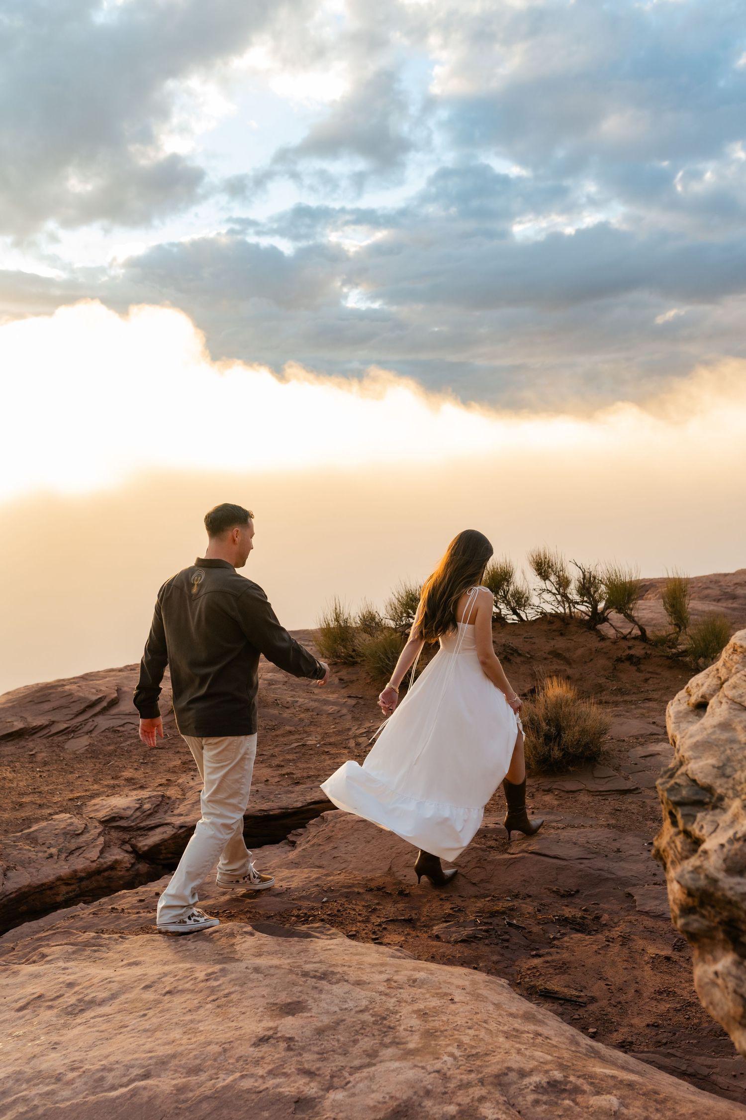 Two figures in white dress and dark jacket stand on red rock formations at sunset against a cloudy sky.