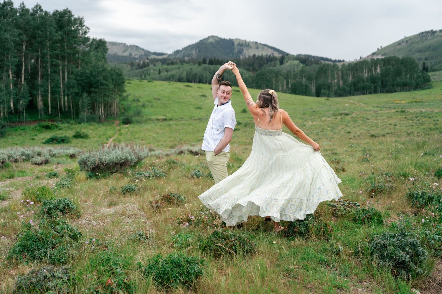 A couple in wedding attire dances joyfully across a scenic mountain meadow with evergreen forests and peaks in the background.