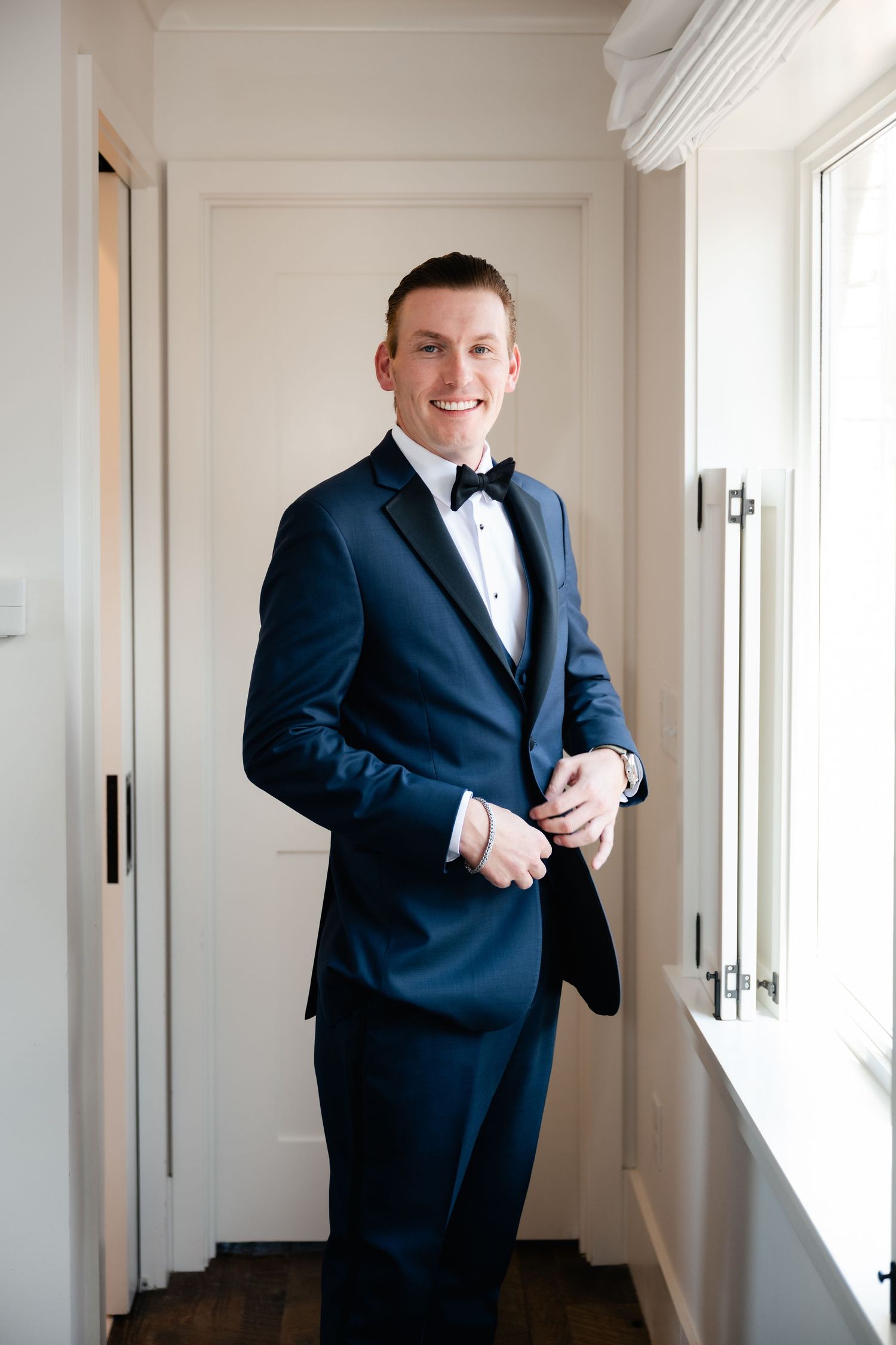 Groom in navy tuxedo and bowtie poses in bright hallway with white walls and windows.