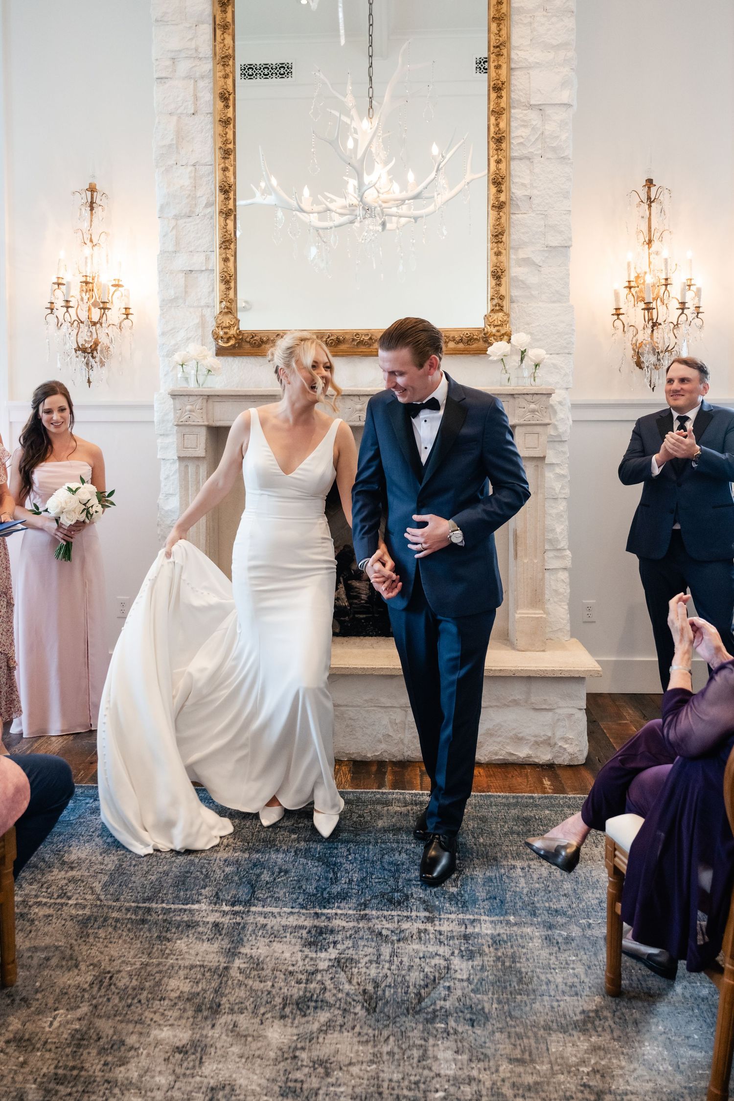 Wedding party members seated in a formal room with white walls and decorative lighting fixtures.