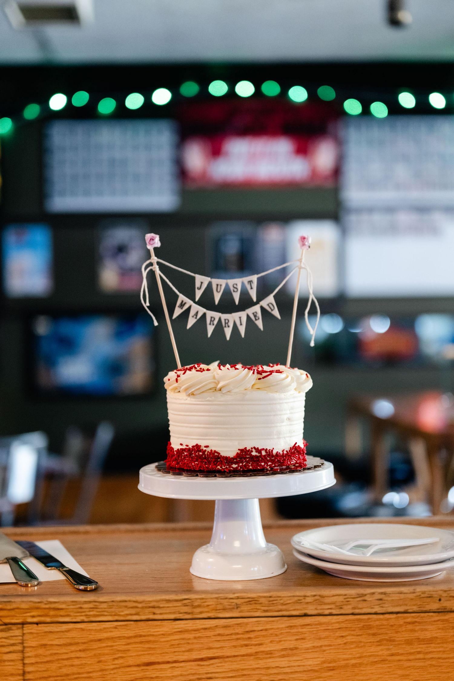 A white wedding cake with bunting topper sits on a wooden table with string lights in background.