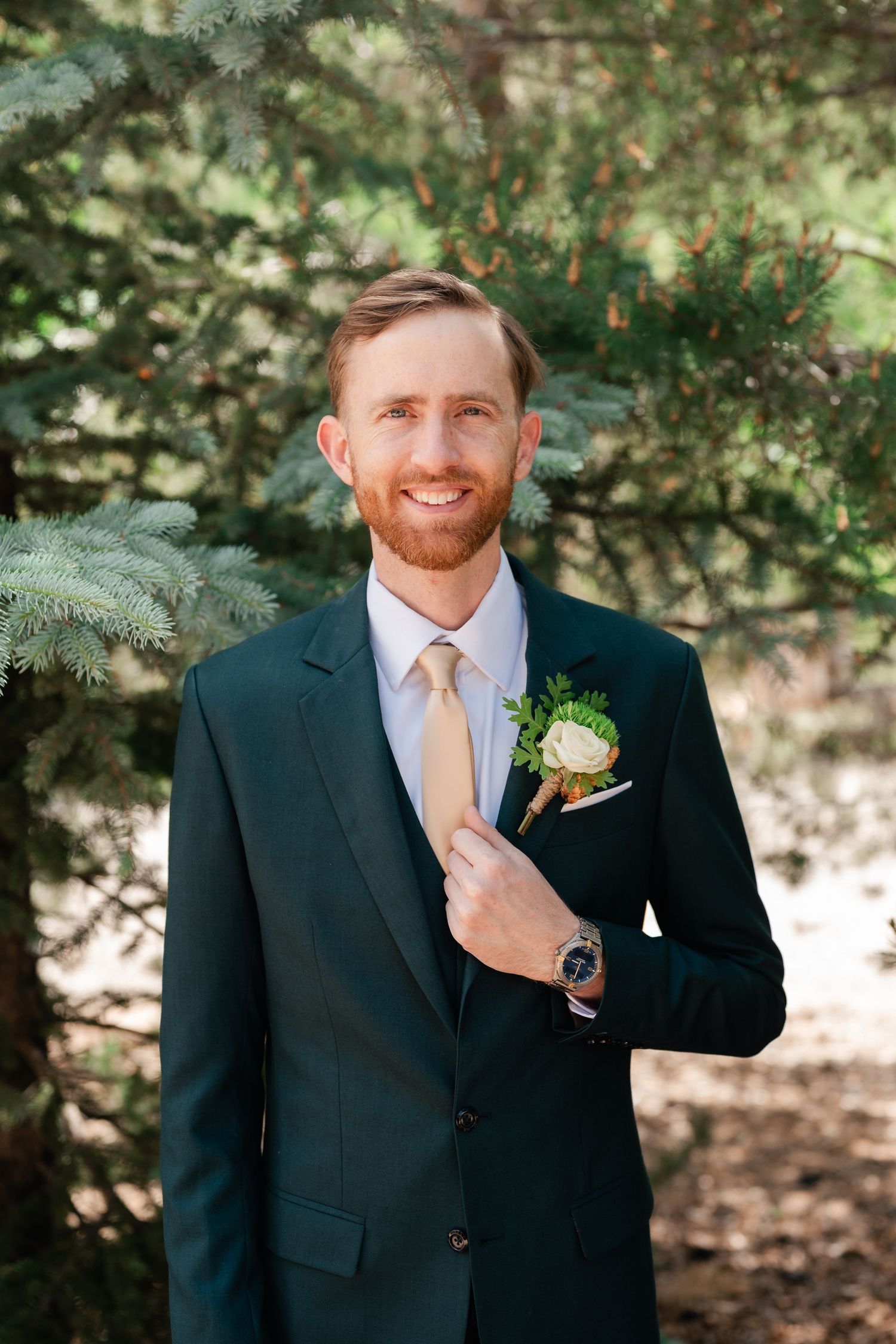 Formal wedding attire featuring a navy suit, yellow tie and boutonniere against evergreen trees.