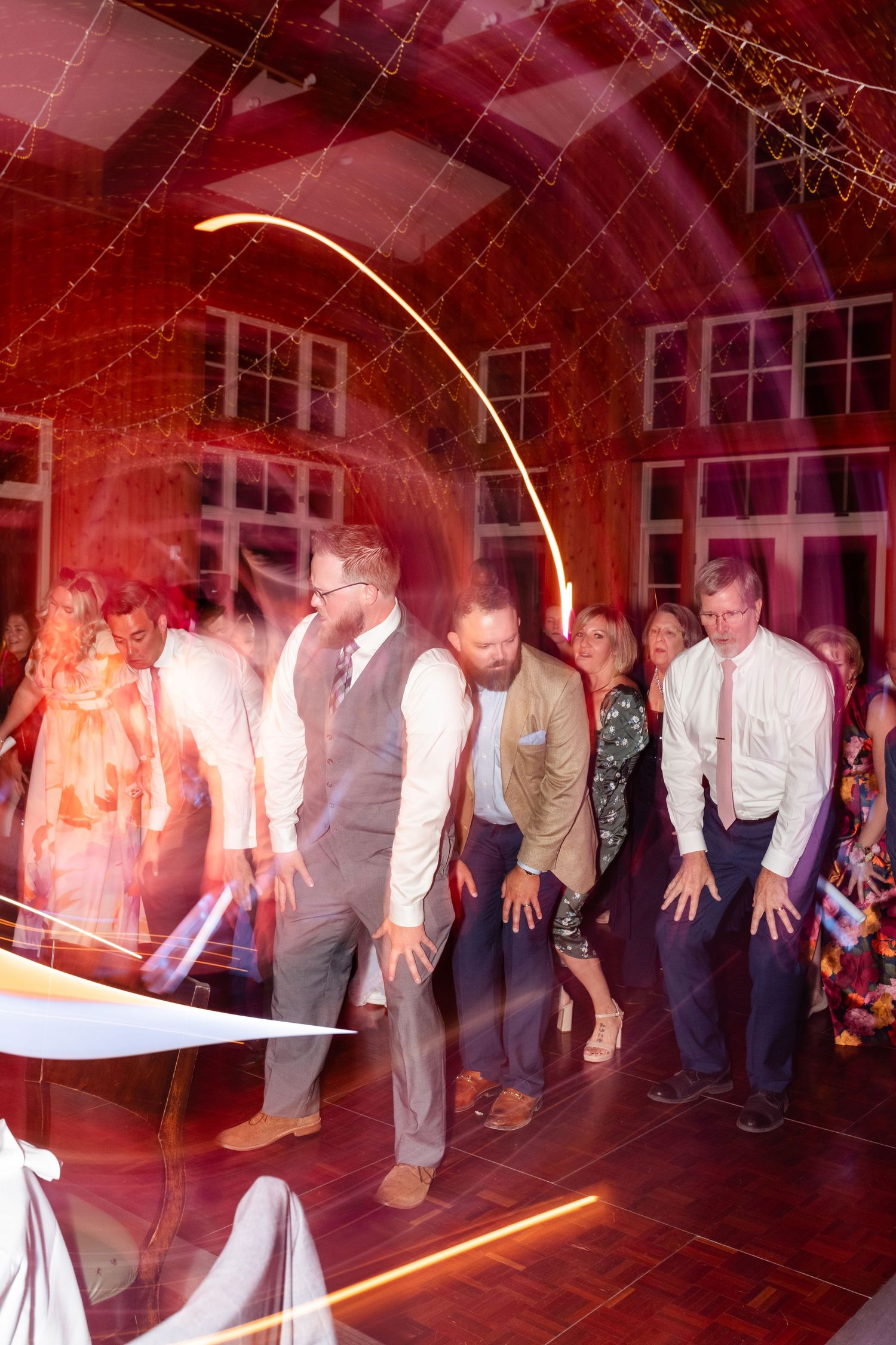 A group of people dancing in a red-lit room with large windows during what appears to be a wedding reception.