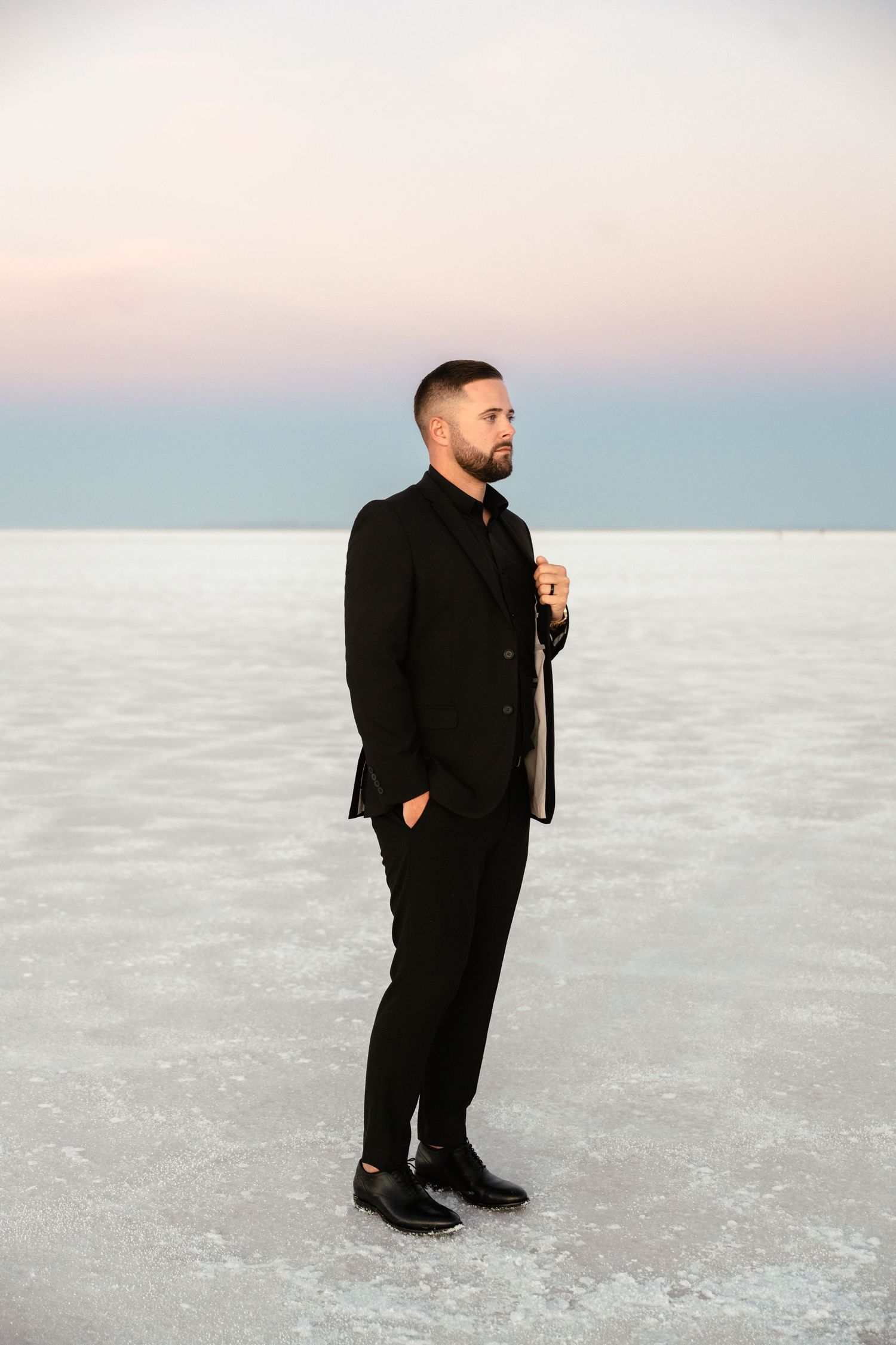 Person in black formal attire standing alone on vast white salt flats at sunset.