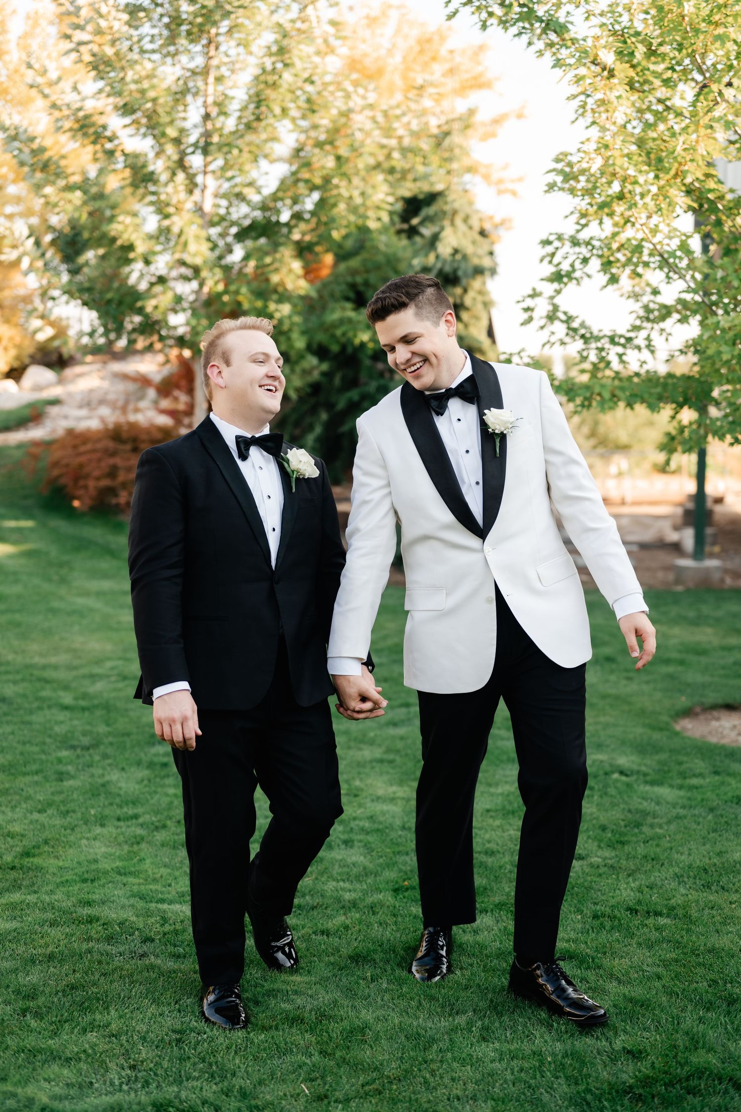 Two grooms in tuxedos walk hand in hand on green lawn, one in white jacket, one in black, both wearing boutonnieres.