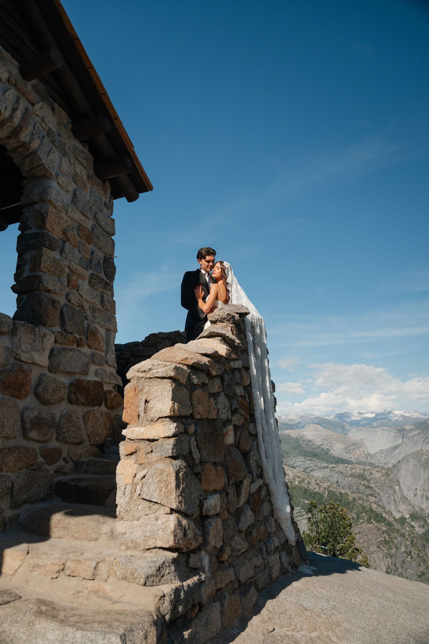 Bride and groom embrace atop a stone wall at Glacier Point, Yosemite, with mountain panorama and blue sky backdrop.