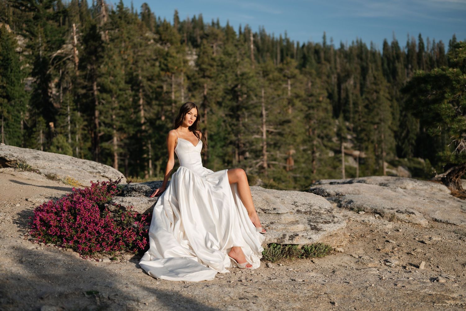 Bride in white wedding gown posing on rocky terrain with pink wildflowers and pine forest backdrop.