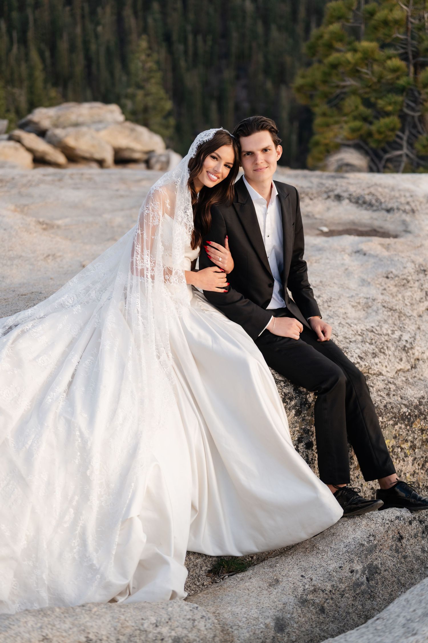 Bride in white lace gown and groom in black suit sit on rocks with pine forest backdrop.