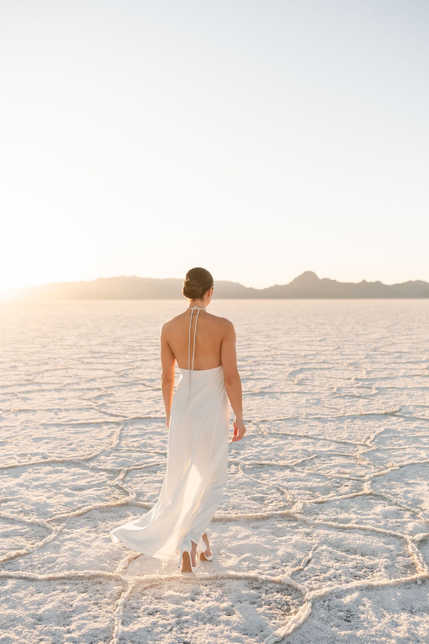 Salt Flats Photographer. A figure in a flowing white dress walks across the stark white salt flats at sunset.