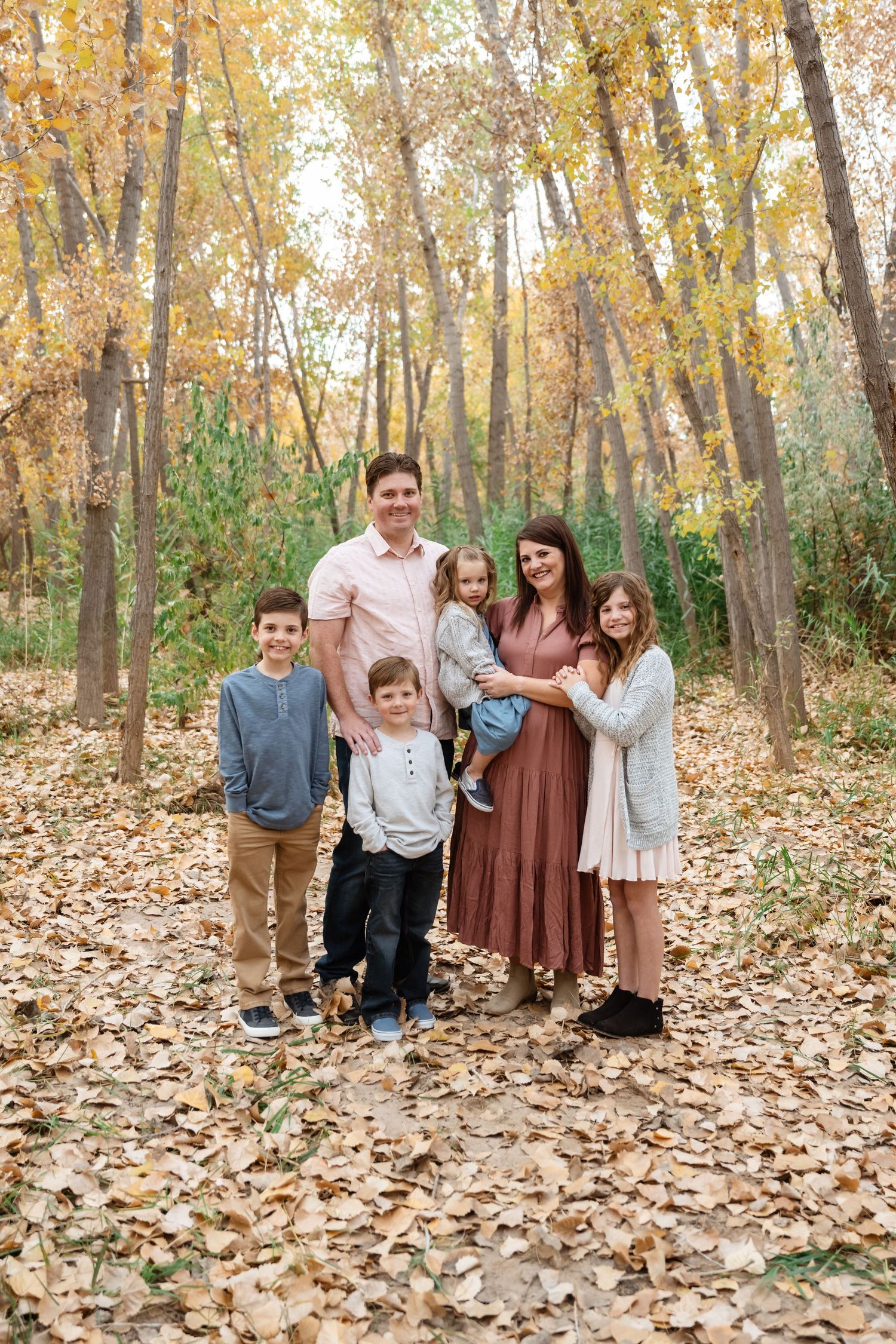 A family poses playfully among autumn trees with fallen leaves covering the forest floor in warm golden light.