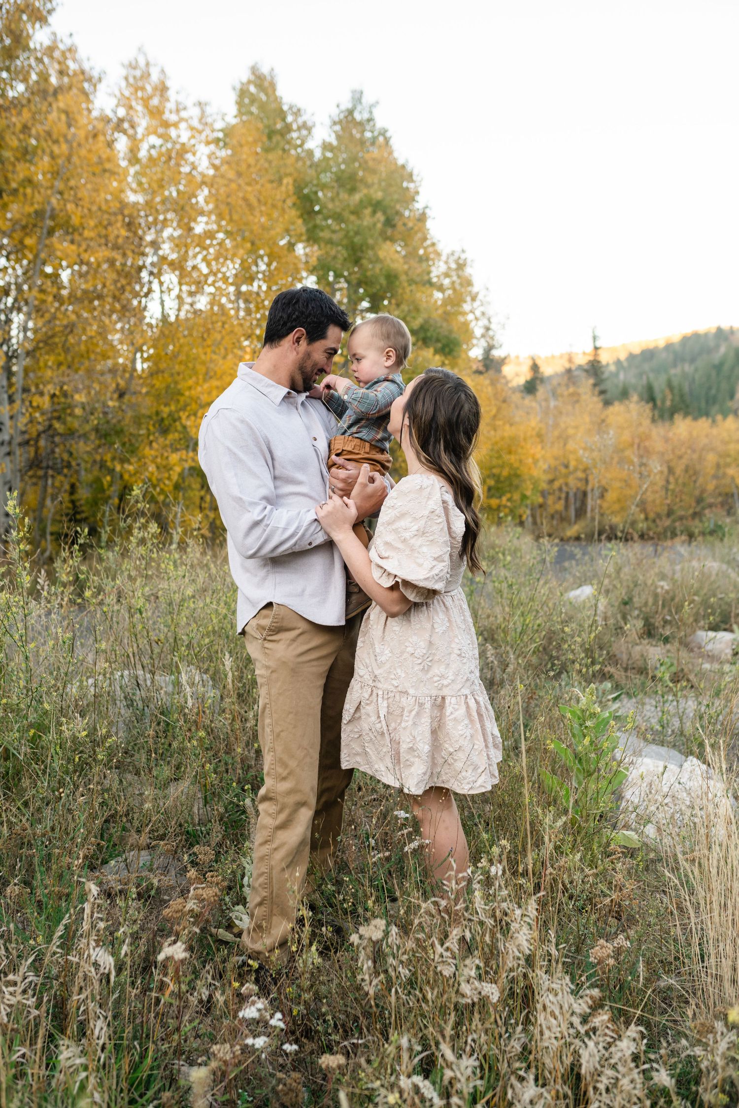 A couple shares romantic moments in a field of tall grass and wildflowers during autumn with golden trees in background.