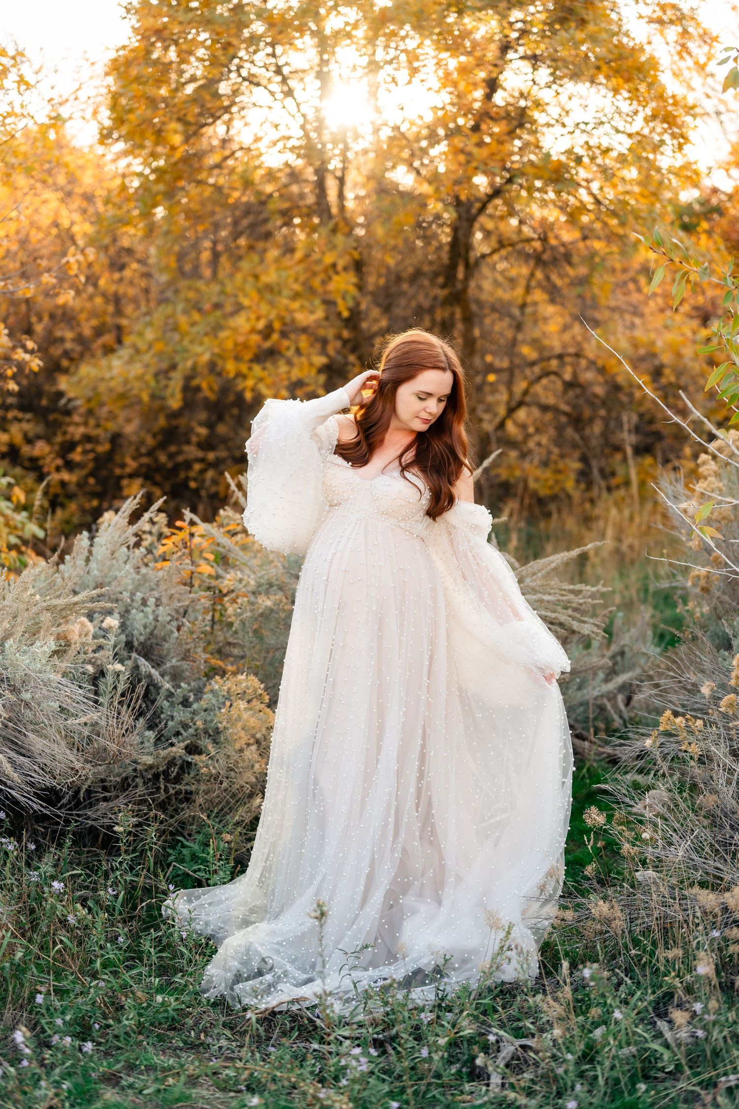 A person in a flowing white lace dress sits gracefully among autumn foliage and tall grasses at golden hour.
