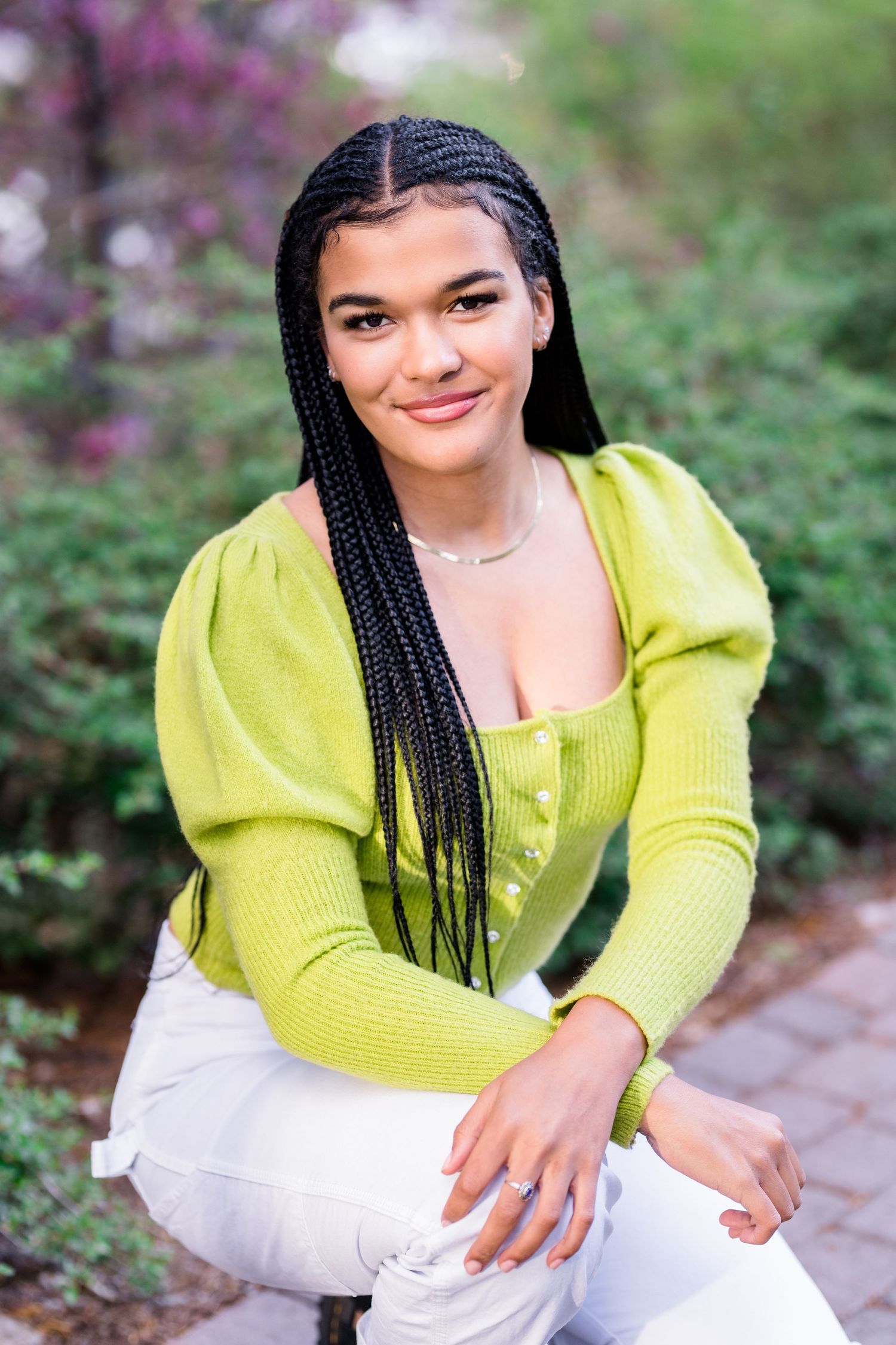 A person in a bright yellow sweater and white pants poses outdoors against a natural background with purple flowers.