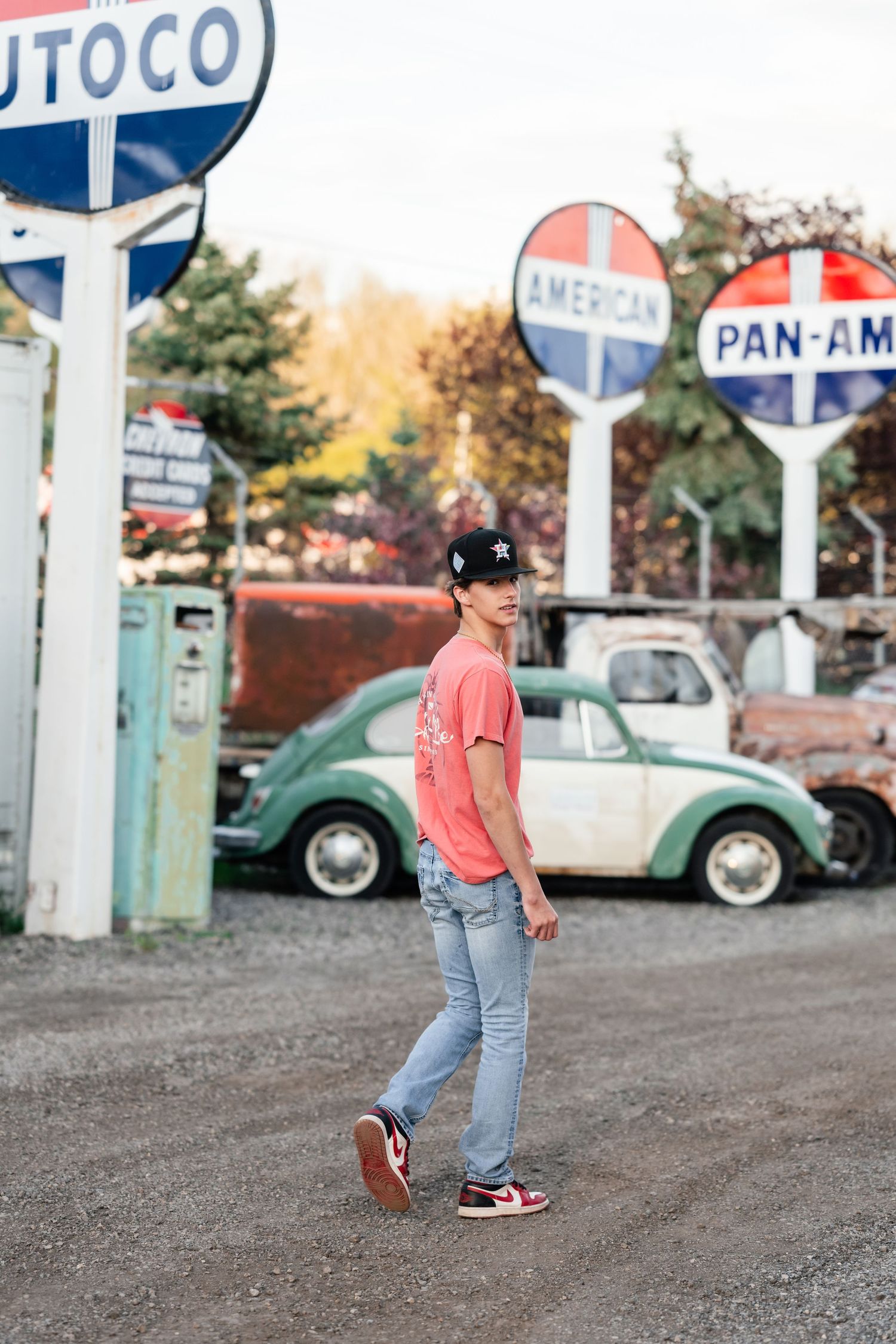 A person in jeans and pink shirt walks on gravel near vintage gas station signs and classic cars in background.