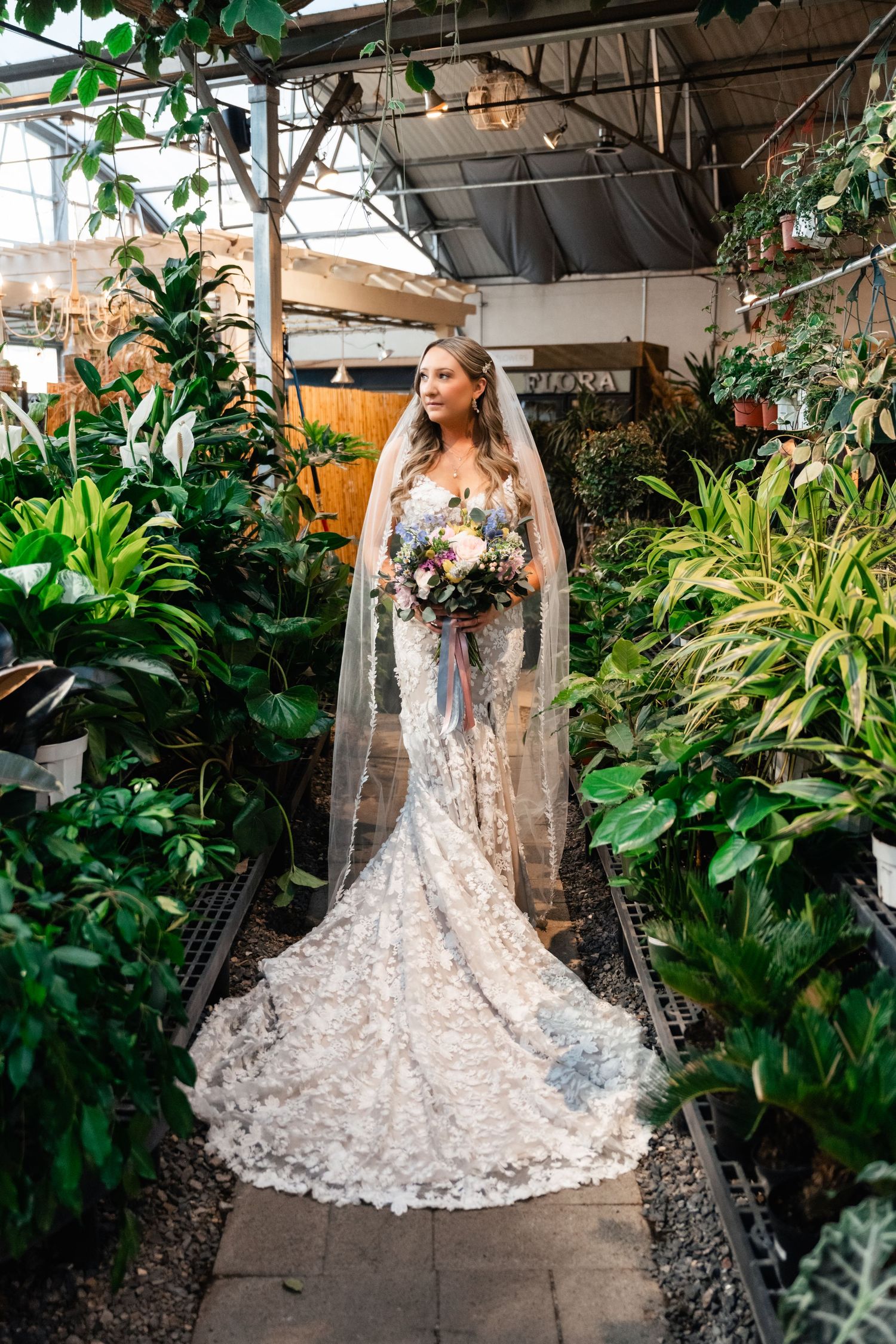 A stunning lace wedding gown with flowing train surrounded by lush greenery in a garden greenhouse setting.