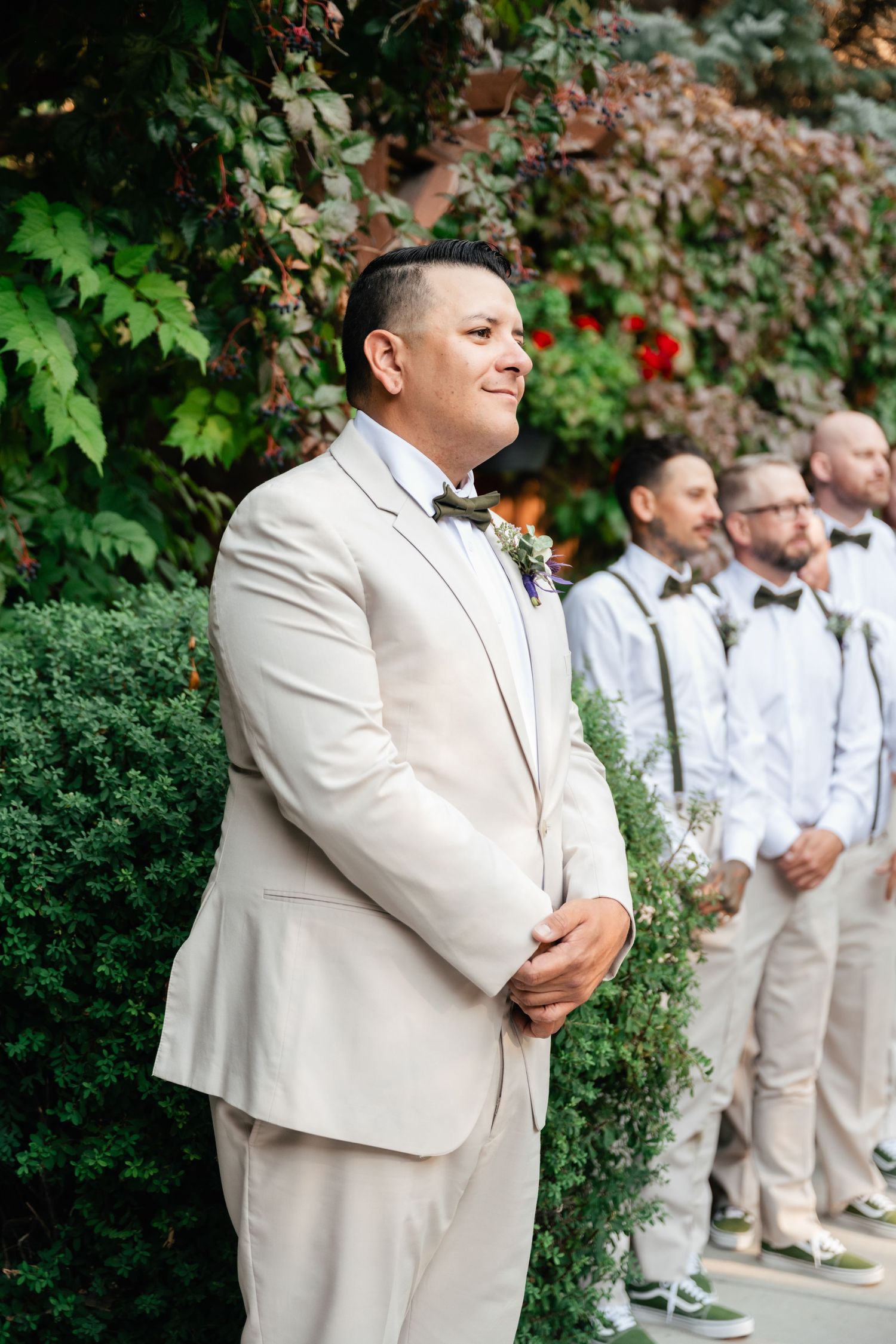 Person in cream-colored suit standing with wedding party against lush garden backdrop.