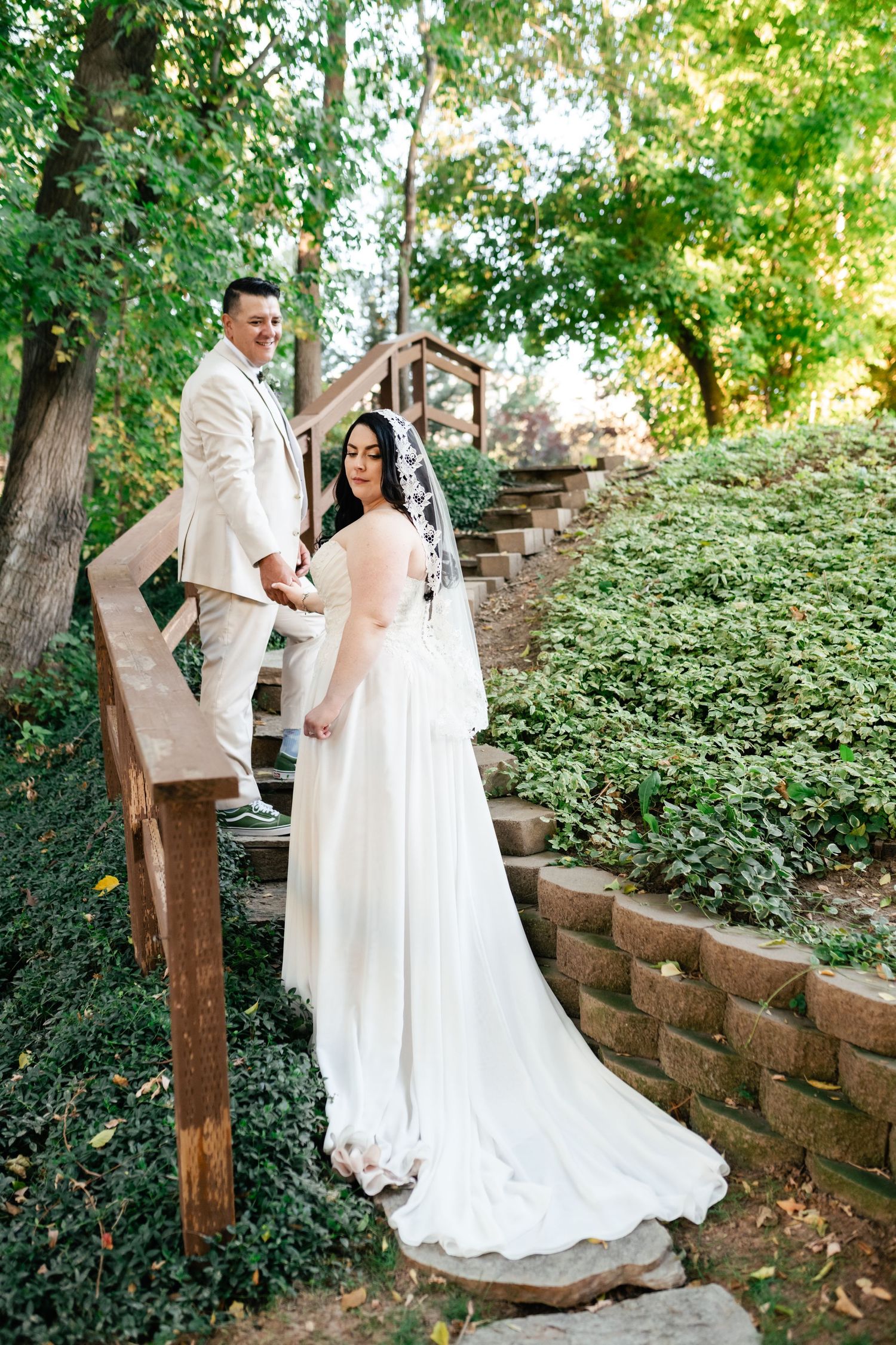 A couple in flowing white attire poses on rustic garden steps surrounded by lush greenery on their wedding day.