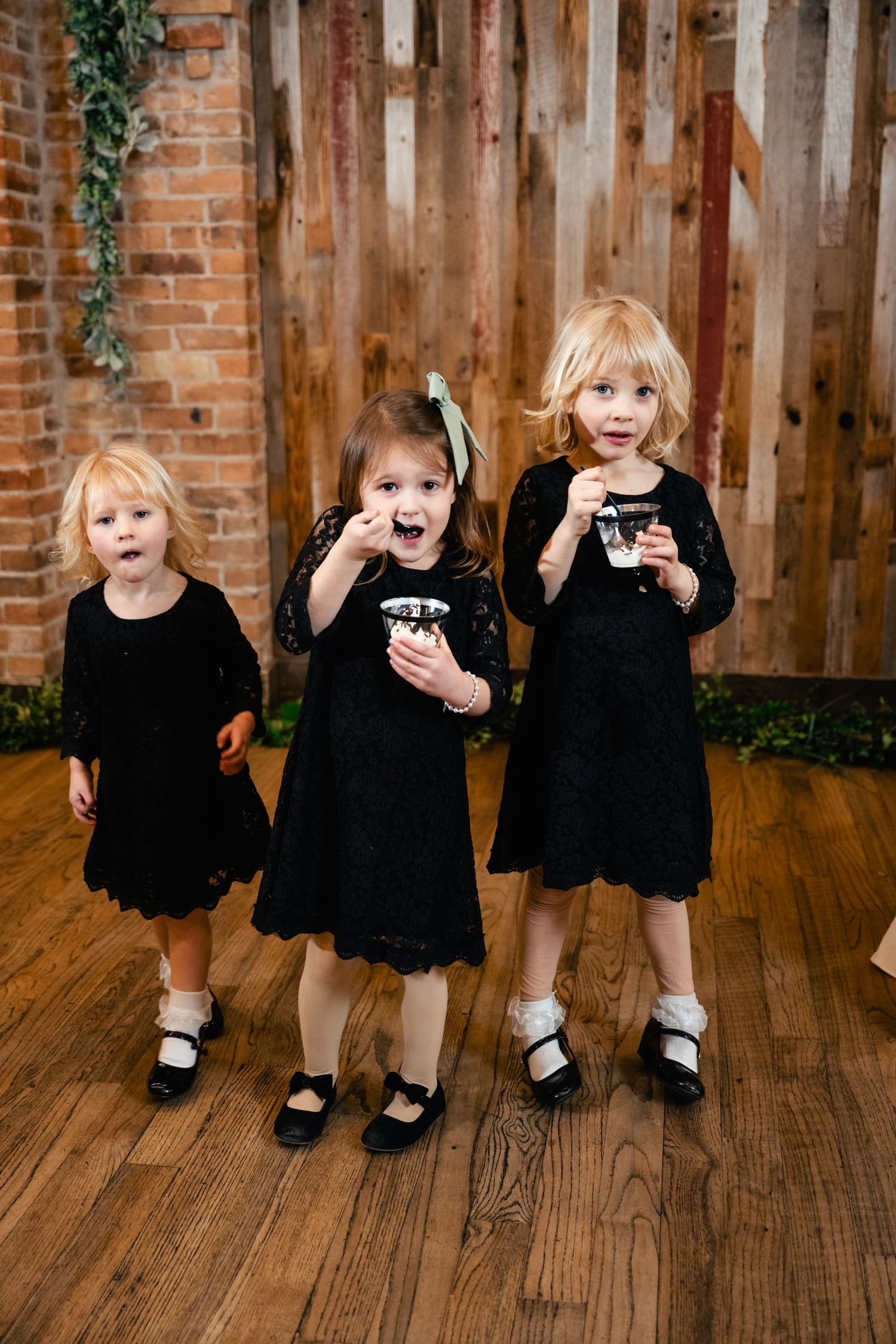 Three children in matching black dresses enjoying cupcakes against a rustic wood and brick wall backdrop.