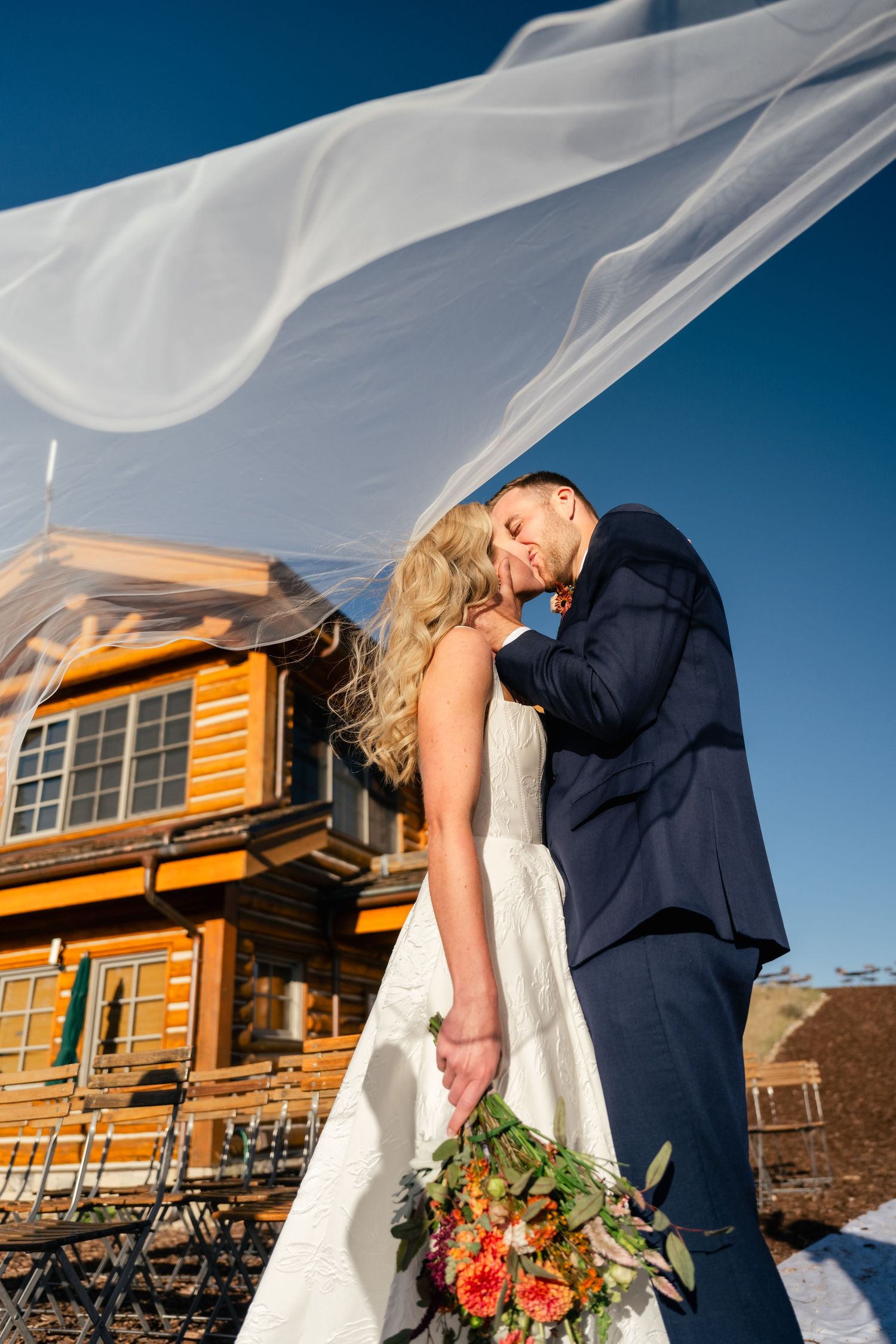 Bride and groom share a kiss beneath flowing veil with rustic wooden lodge in background and dramatic blue sky. Utah Wedding Photographer.