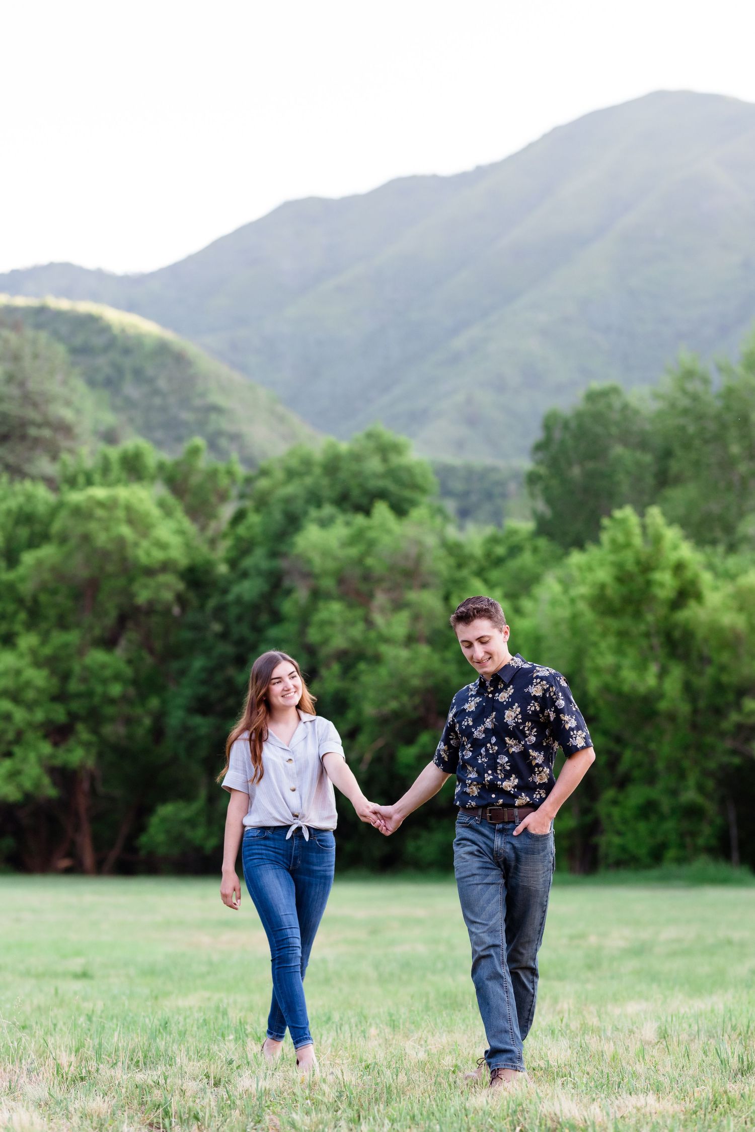 A couple walks hand in hand through a grassy meadow with mountains in the background.