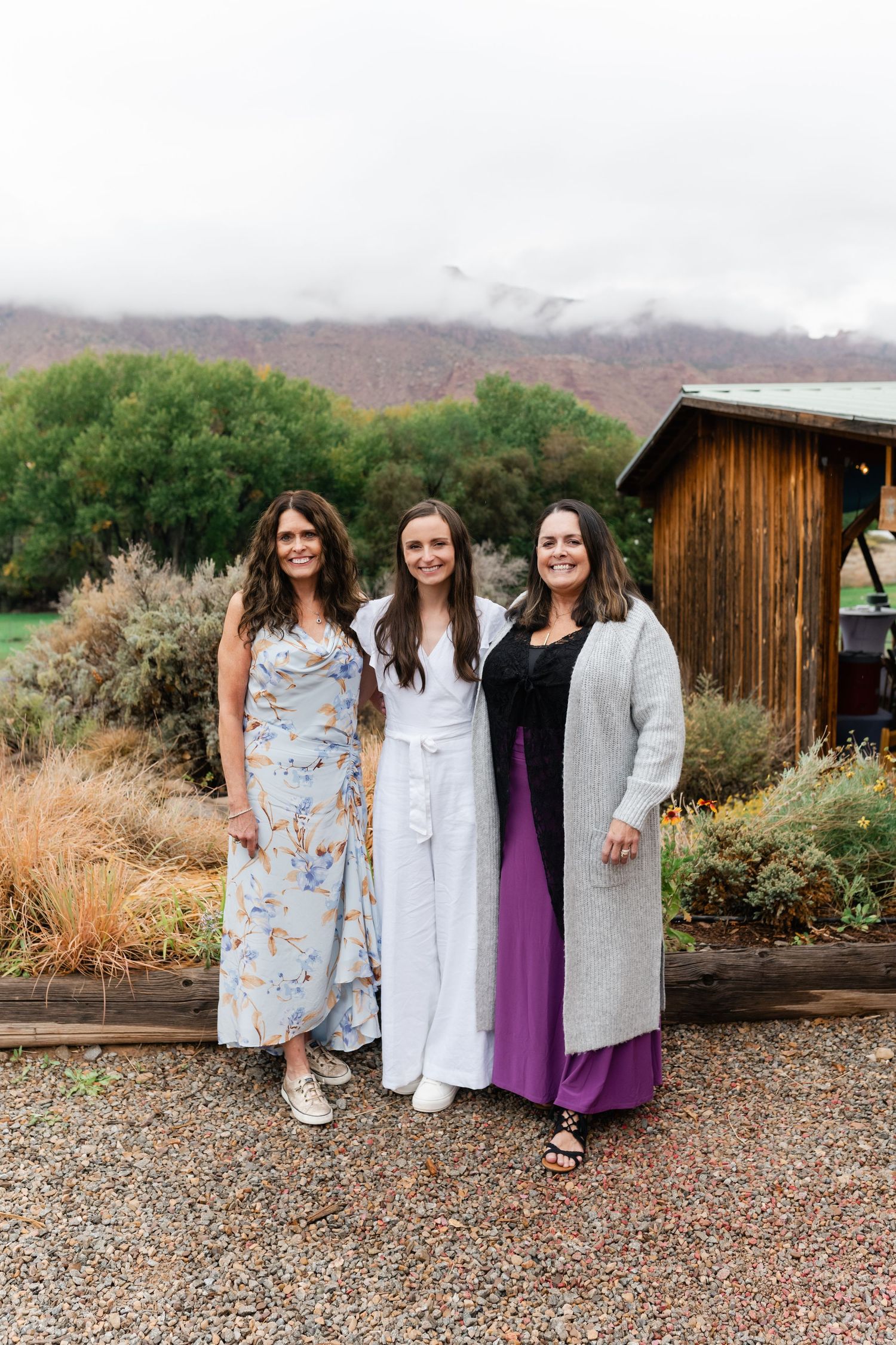 Wedding party members in light colored dresses and gowns pose together on gravel path near desert landscape.